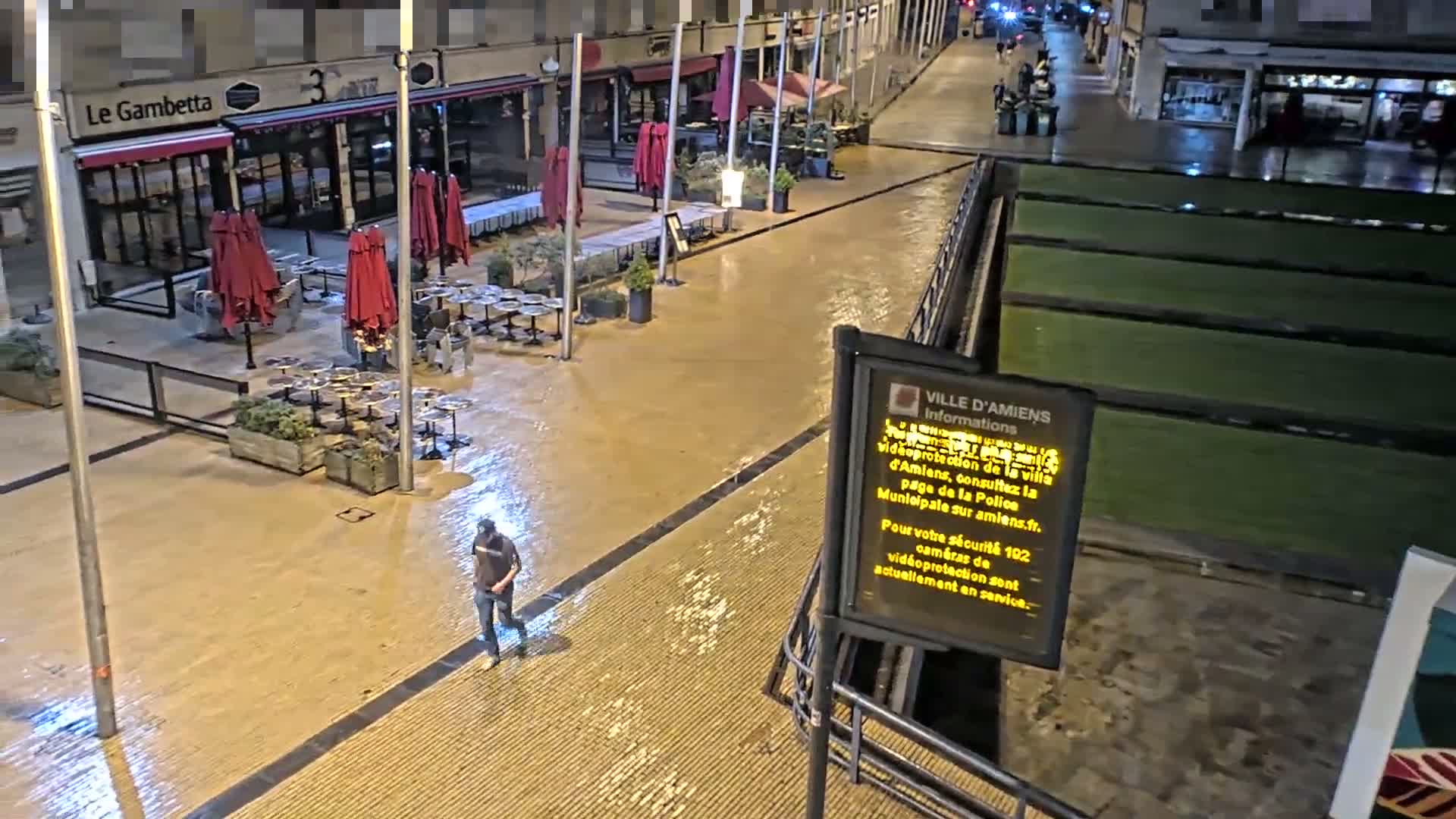 A person walks through a wet, paved plaza at night, past closed outdoor cafes with red umbrellas and adjacent grassy areas.