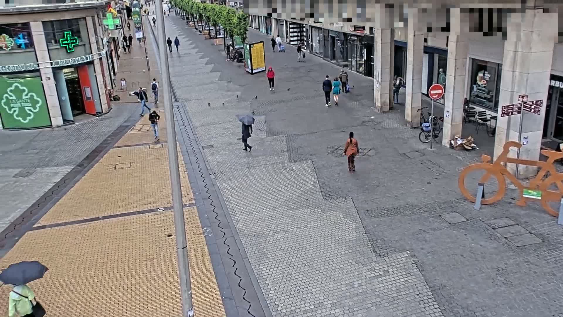 A mostly empty cobblestone pedestrian street is seen on a slightly overcast day, with a few people walking and a person with an umbrella.