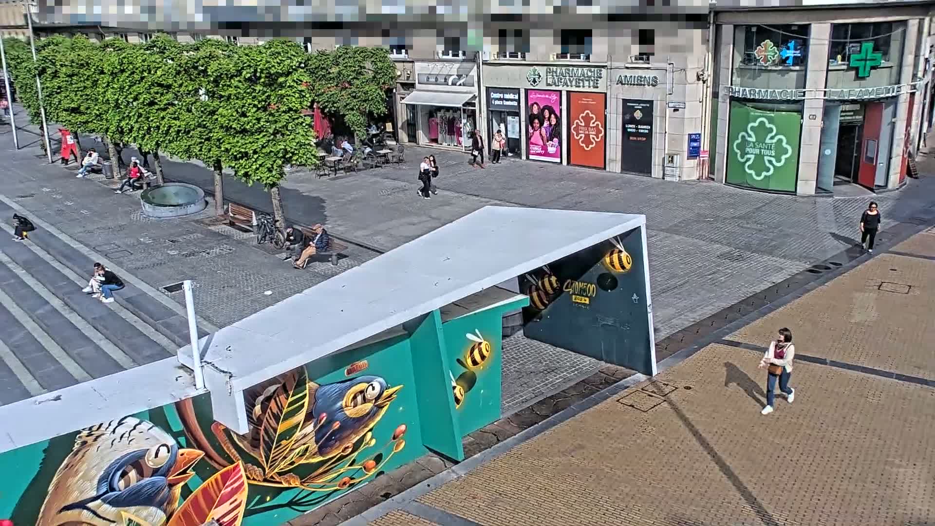 A sunny city square features a mural of birds and bees on a modern structure, with several people sitting and walking around shops and trees.
