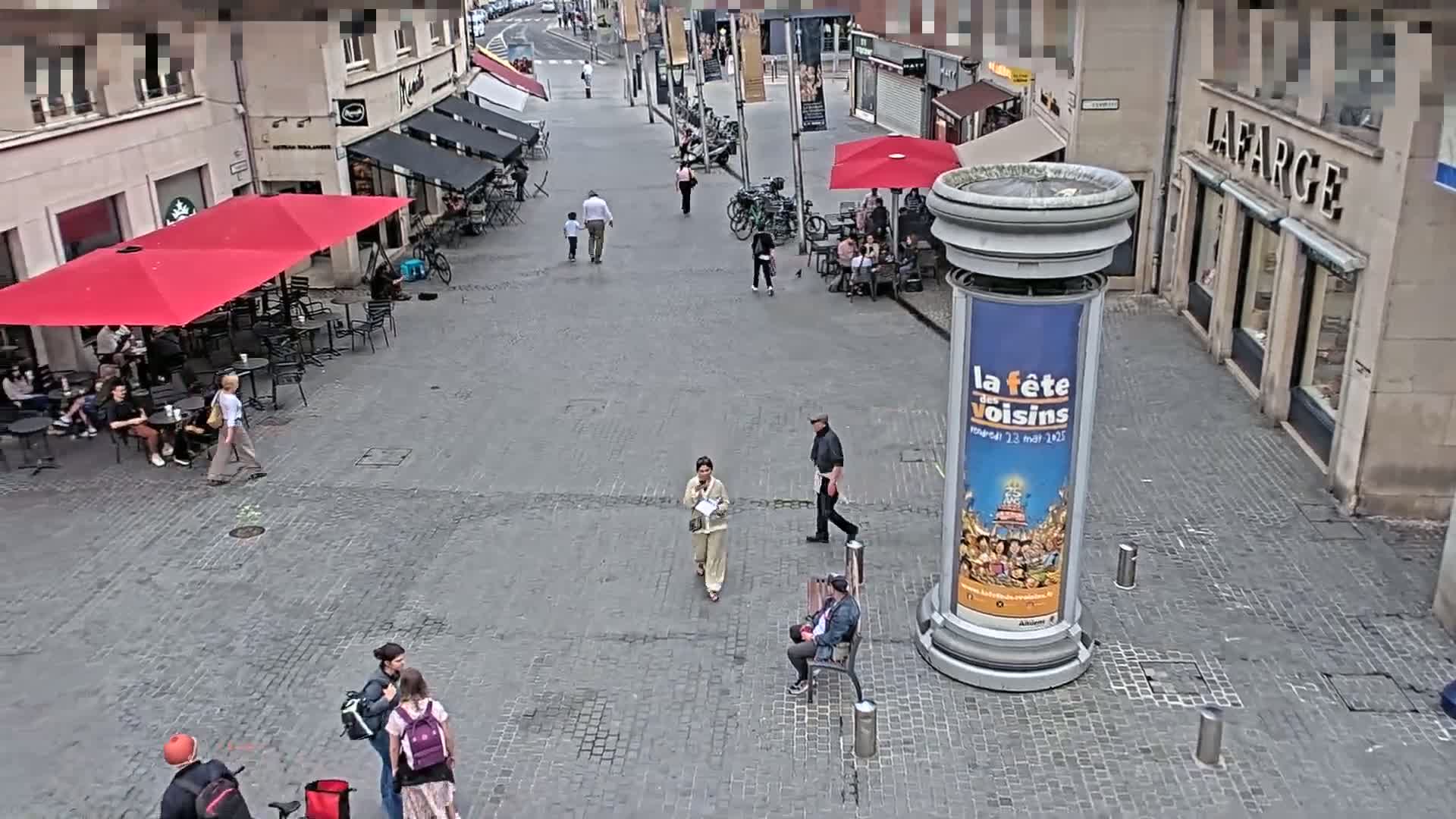 A cobblestone town square, under a partly cloudy sky, features several people walking and sitting at outdoor cafes under red umbrellas.