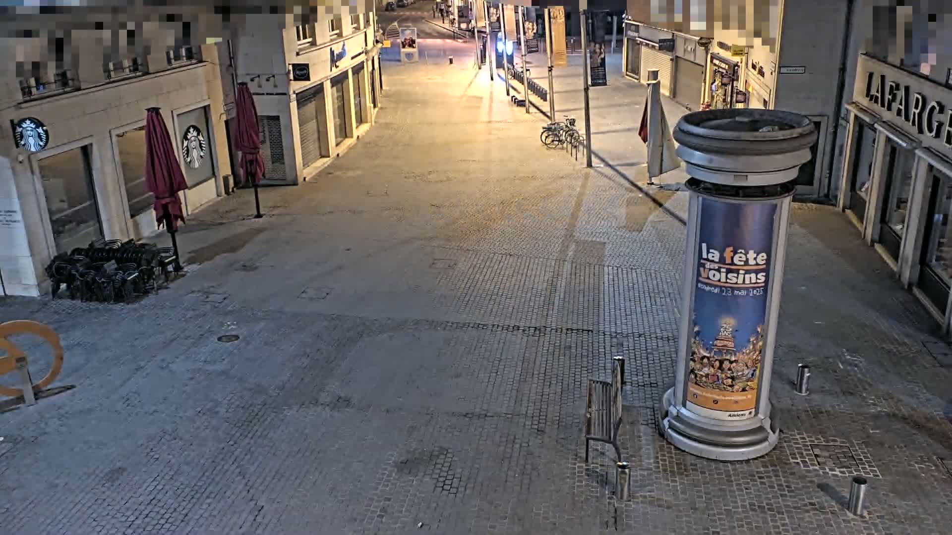 A mostly empty, cobblestone street at night is lit by streetlights, with a few closed shops and a single bench visible.