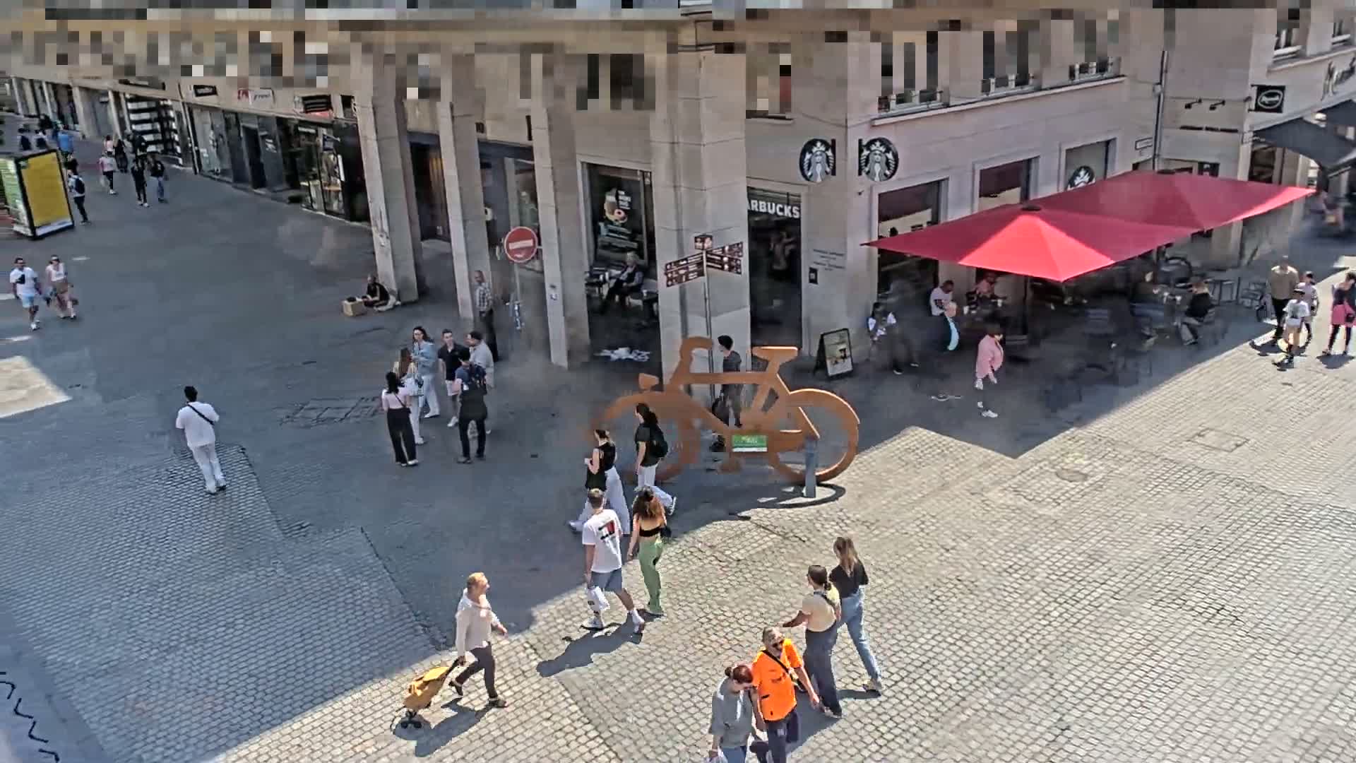 A paved plaza on a sunny day with several people walking around a large bicycle sculpture near a Starbucks.