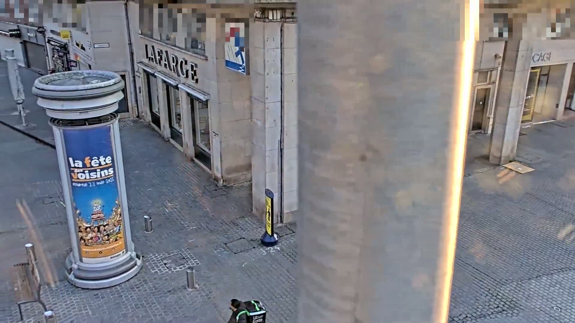 A person carrying a delivery bag walks along a cobblestone street past buildings and a large advertisement column.
