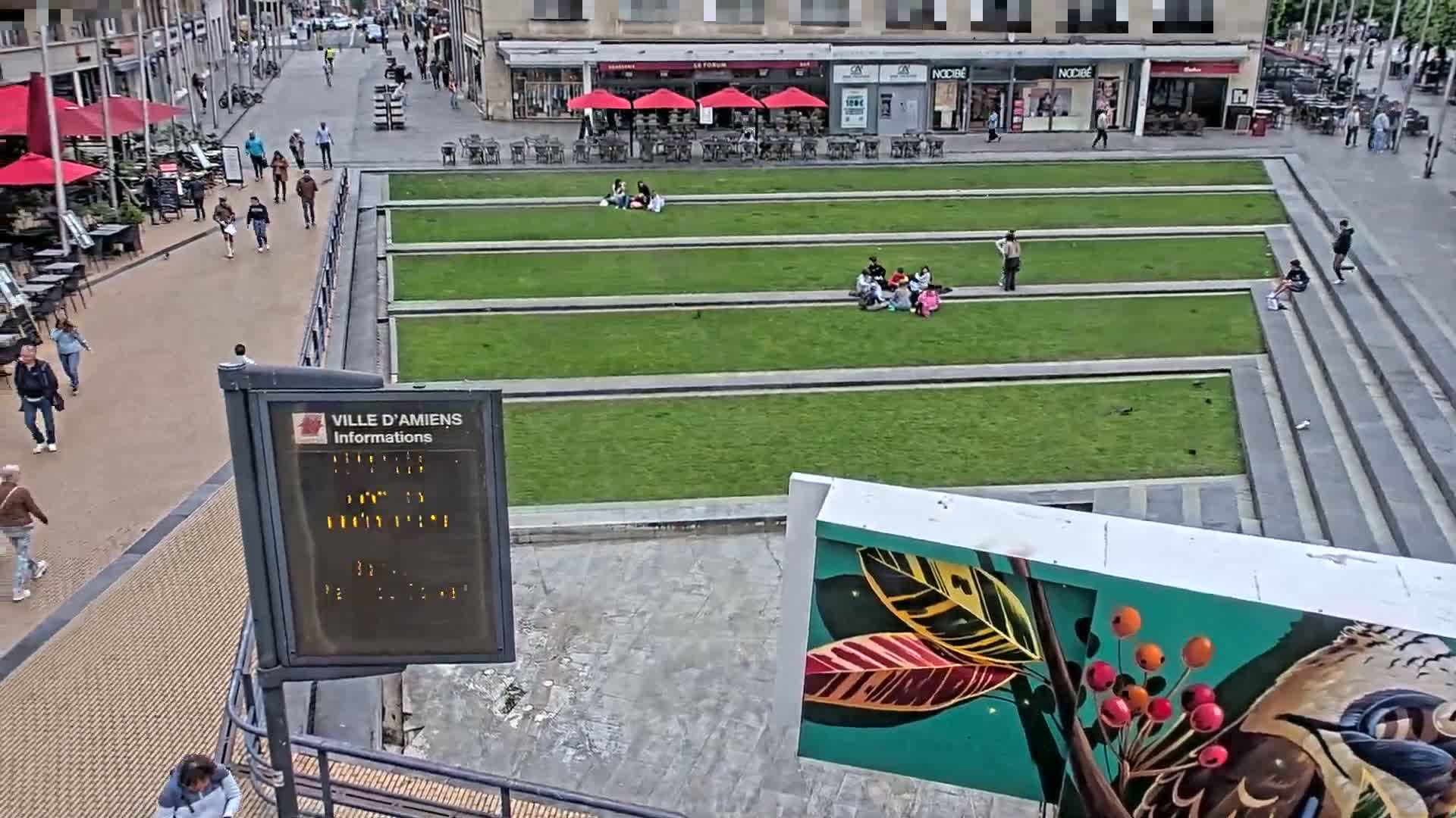 A city square with several groups of people relaxing on grassy areas, under a partly cloudy sky.