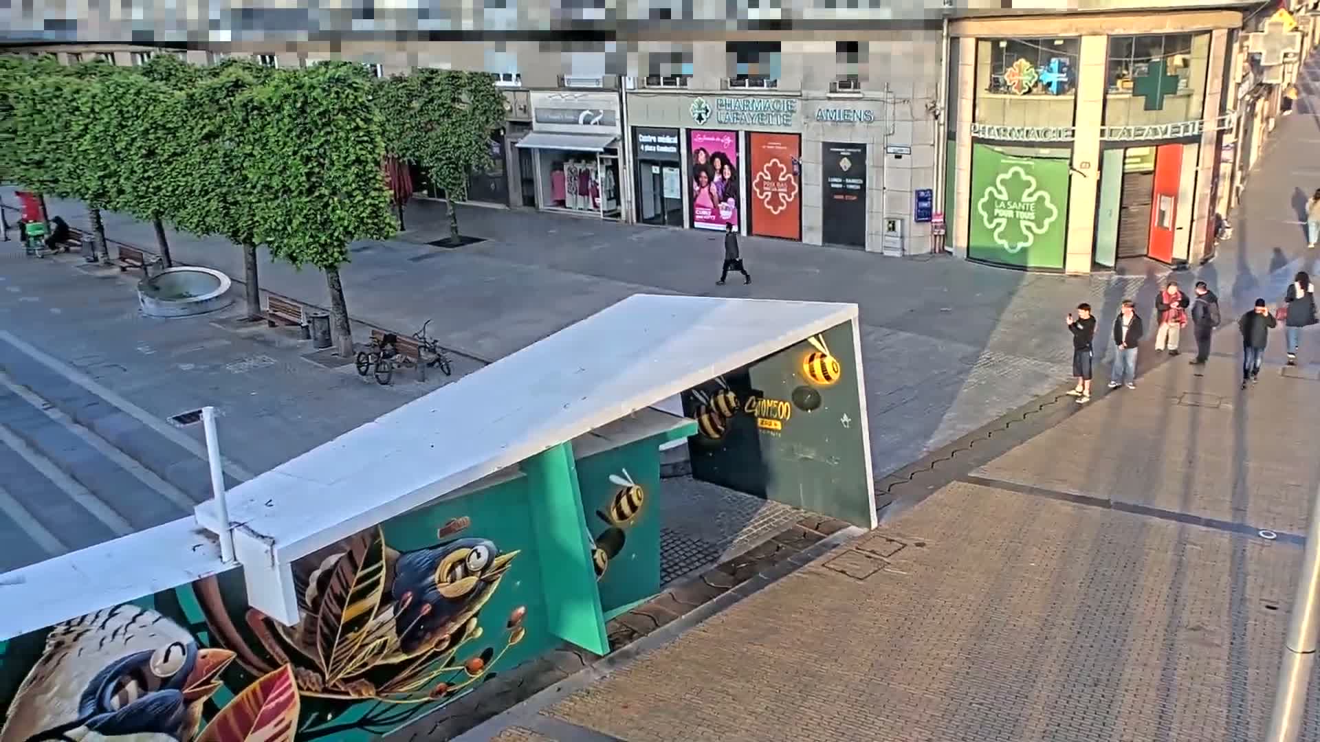 A sunny city street scene shows several pedestrians walking past shops and a uniquely painted structure with a white overhang.