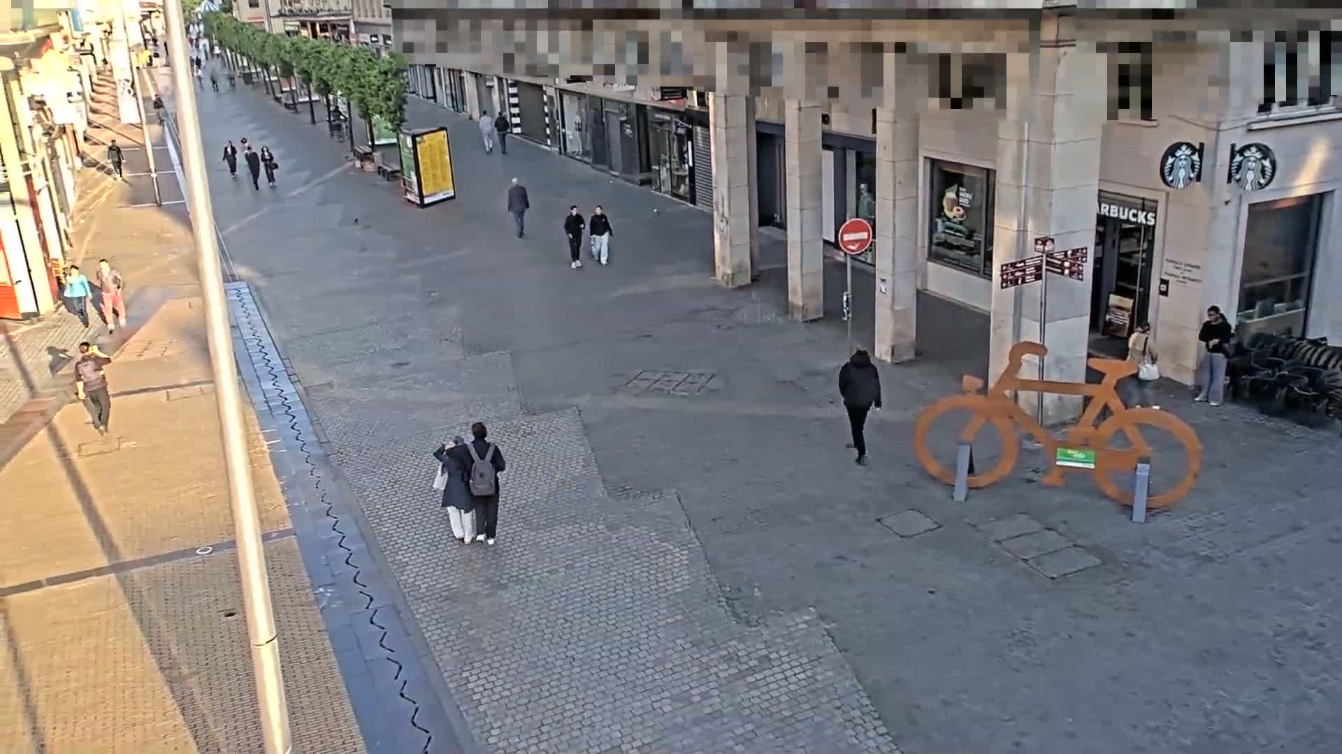 A paved pedestrian area with a few people walking past shops and buildings on a sunny day, featuring a large, decorative bicycle sculpture.
