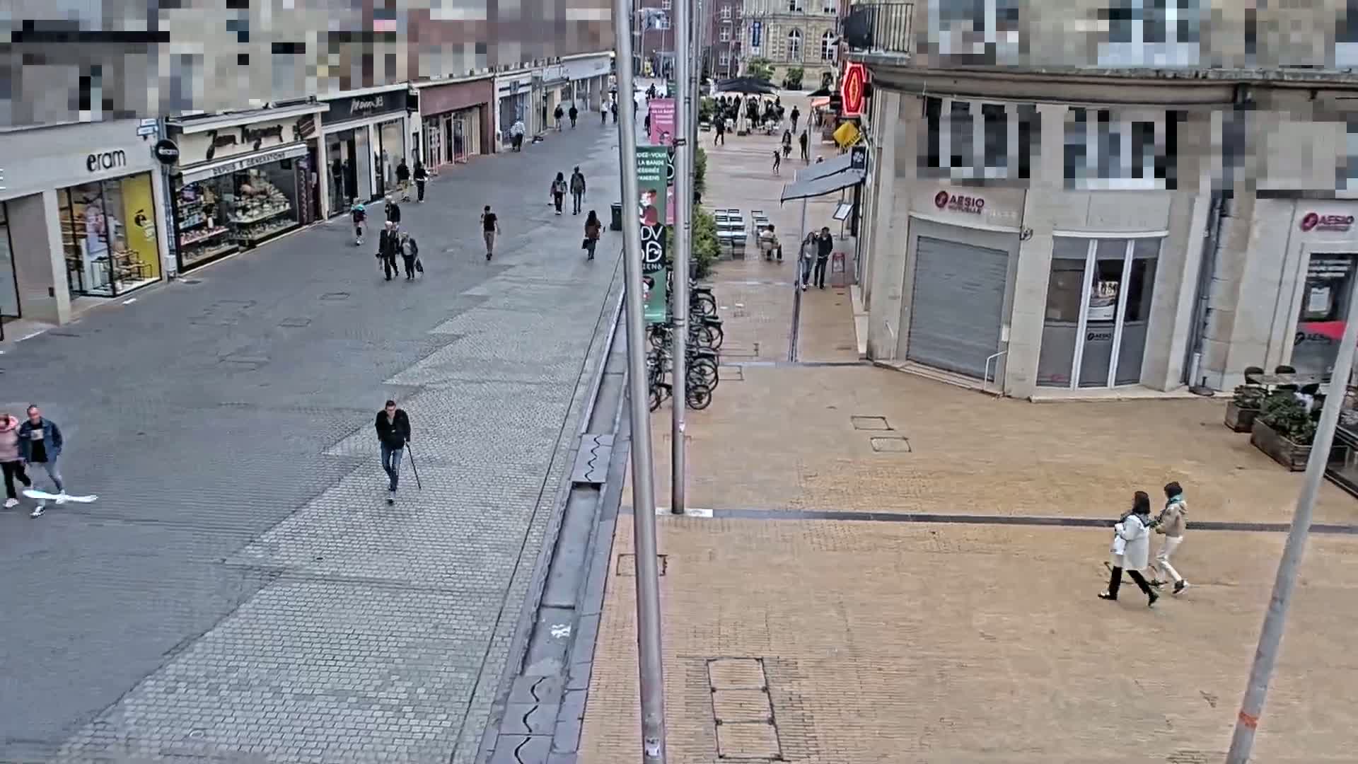 A mostly empty pedestrian street with several people walking, lined with shops and buildings, on a partly cloudy day.