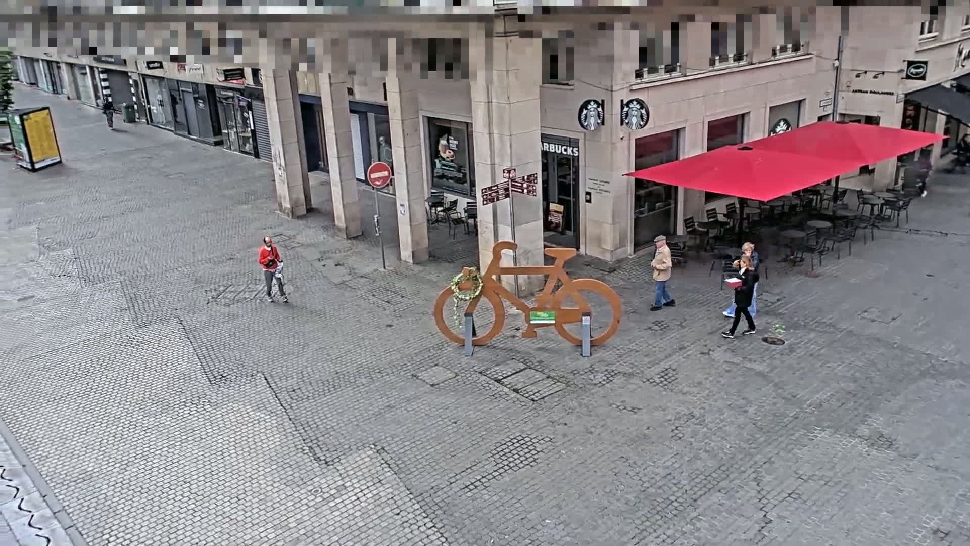A cobblestone city square features a large wooden bicycle sculpture, several pedestrians, and a Starbucks with a red awning under a mostly clear sky.