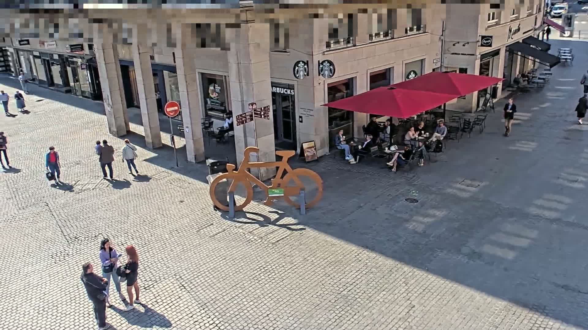 A sunny cobblestone plaza shows people walking and sitting at outdoor tables under red umbrellas outside a Starbucks.