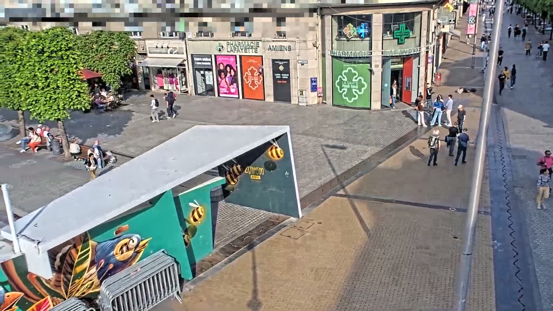 A sunny day reveals a city square with several people walking and sitting, alongside shops and a uniquely designed structure with bee artwork.