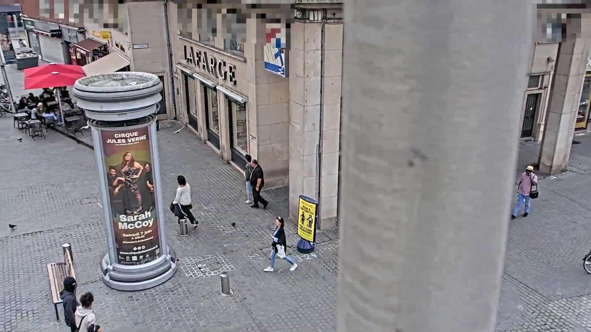A high-angle view of a city street shows several people walking past a large advertisement on a column under a partly sunny sky.