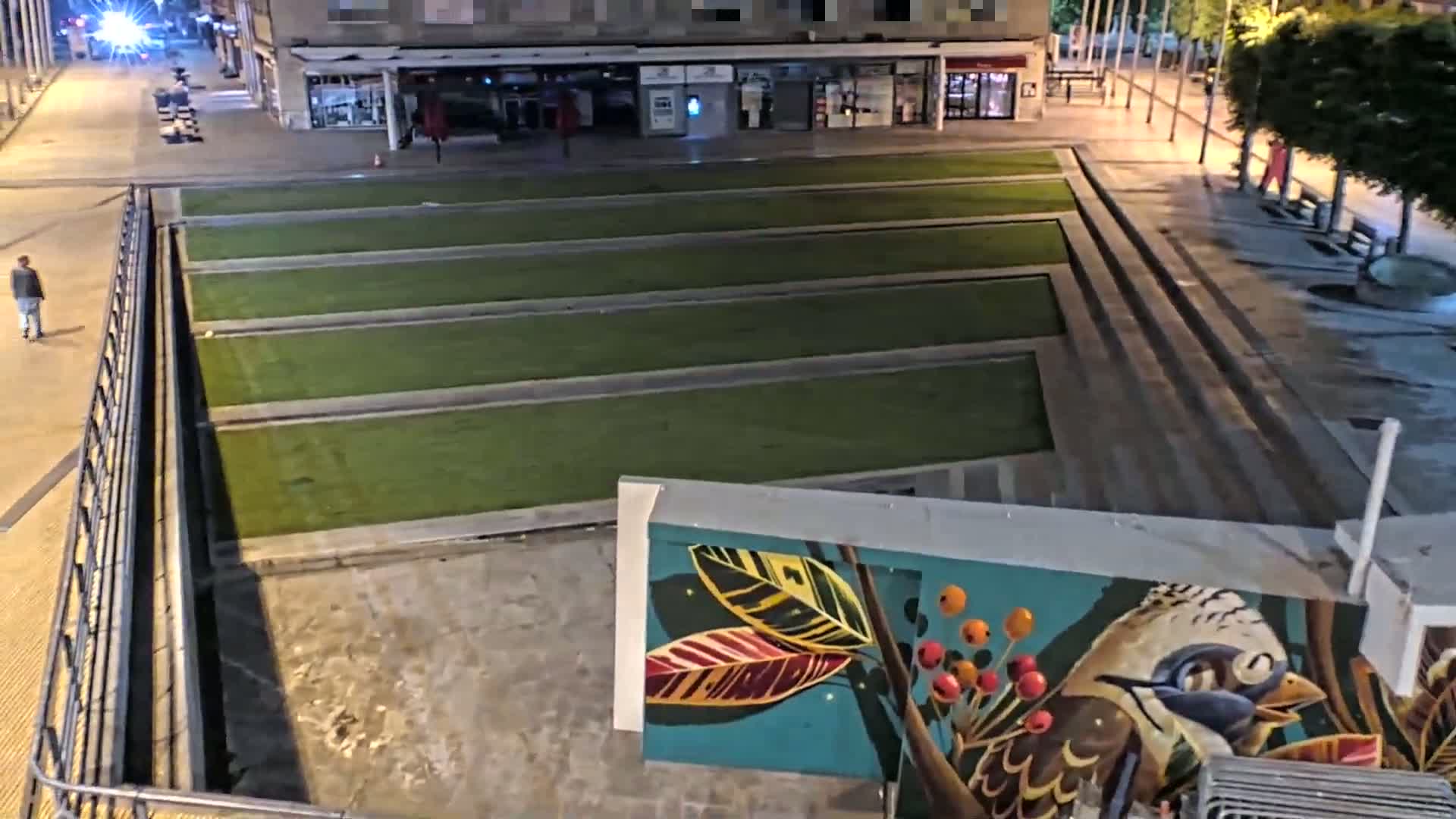 A nighttime, high-angle view shows a paved plaza with rectangular patches of grass, a mural depicting a bird and foliage, and a person walking on a sidewalk.