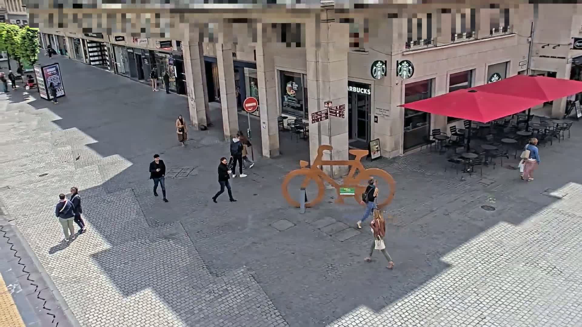 A sunny city street scene shows several pedestrians walking past a Starbucks with outdoor seating and a large, decorative bicycle sculpture.
