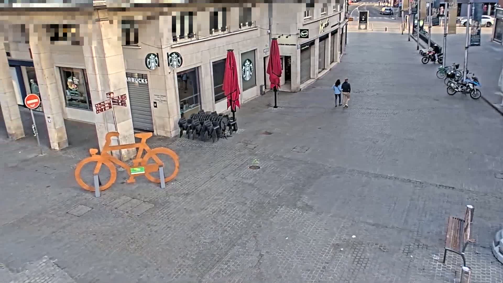 A paved city square with a large orange bicycle sculpture, stacked chairs, two red umbrellas, and a couple walking past a Starbucks.
