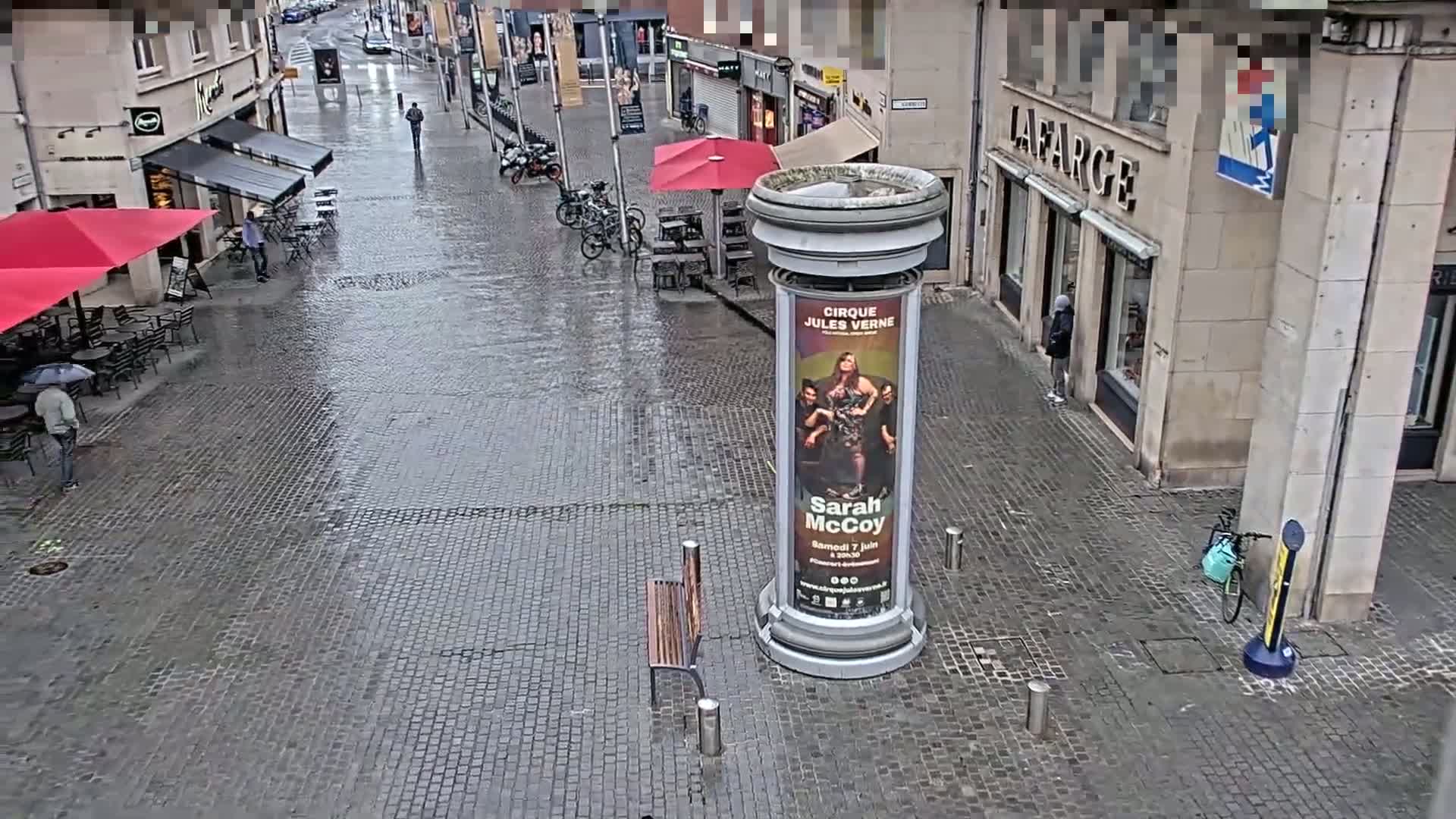 A wet cobblestone street is seen with several people walking past outdoor seating areas under red umbrellas, a bench, and a tall cylindrical advertisement stand.