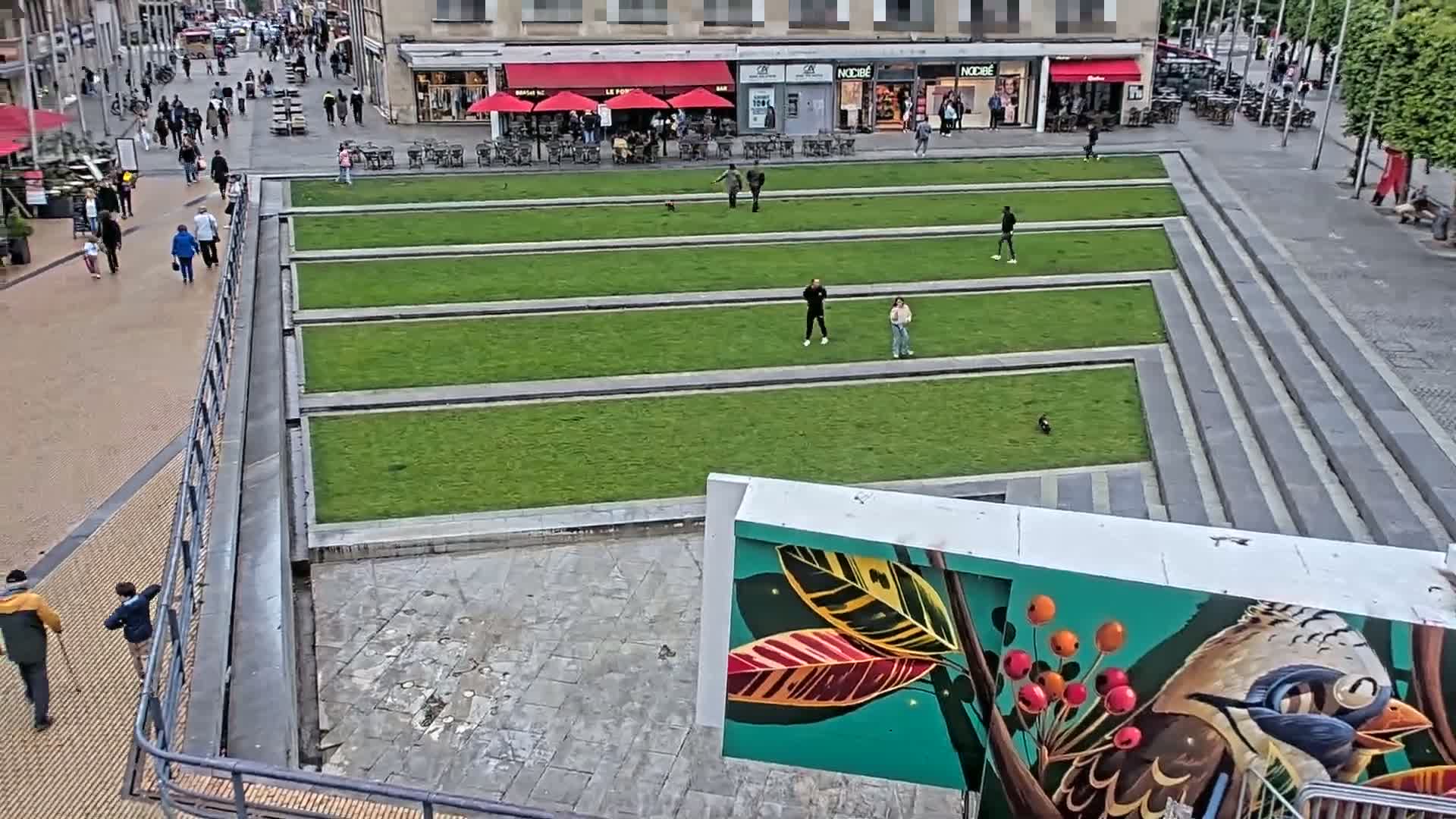 A high-angle view shows a paved city square with rectangular grassy areas, a few pedestrians, and a large mural of a bird on a wall.