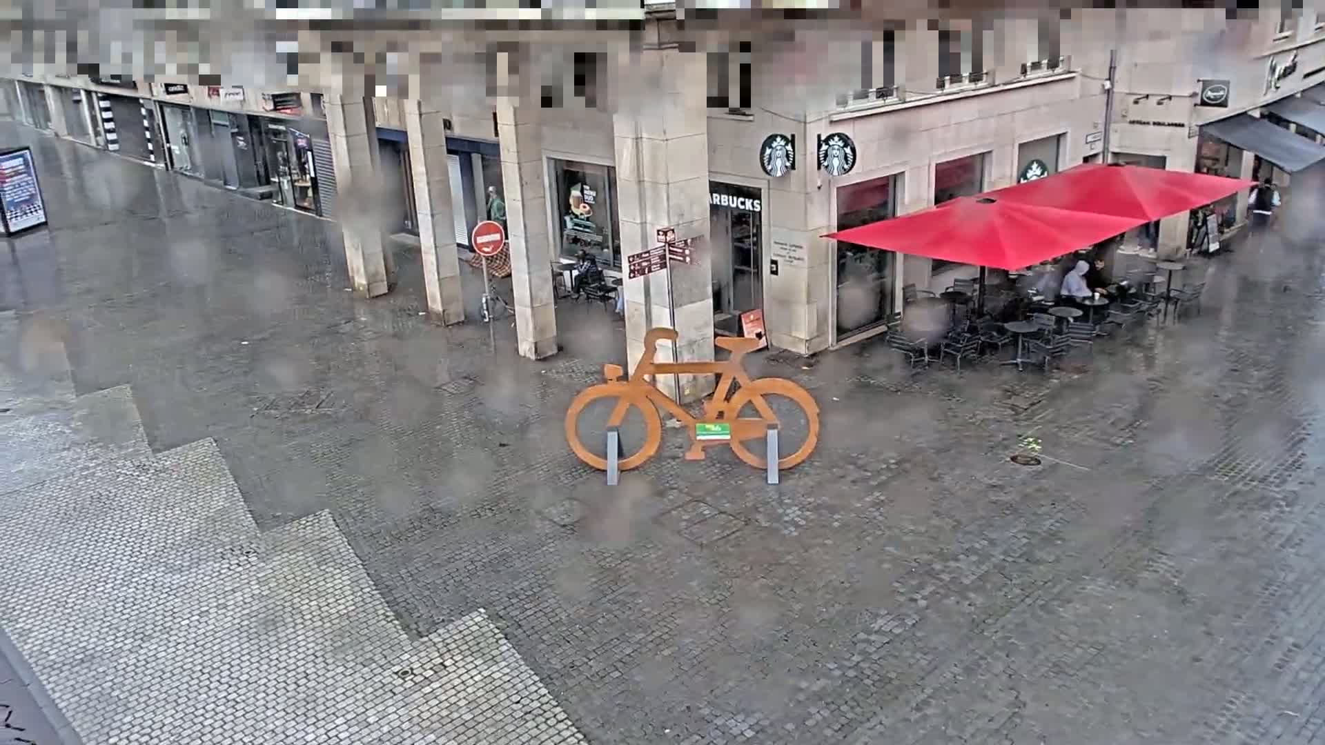 A rain-slicked cobblestone street shows a large, metal bicycle sculpture near a Starbucks with an outdoor seating area under a red umbrella.
