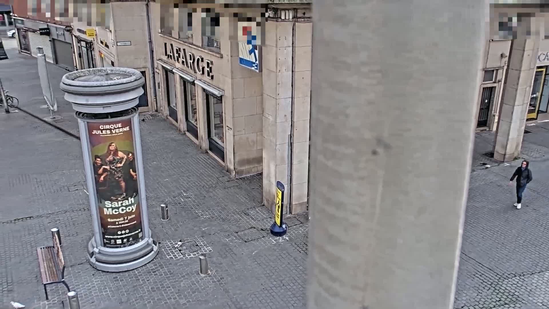 A person walks down a cobblestone street past a column displaying a poster, near buildings on an overcast day.