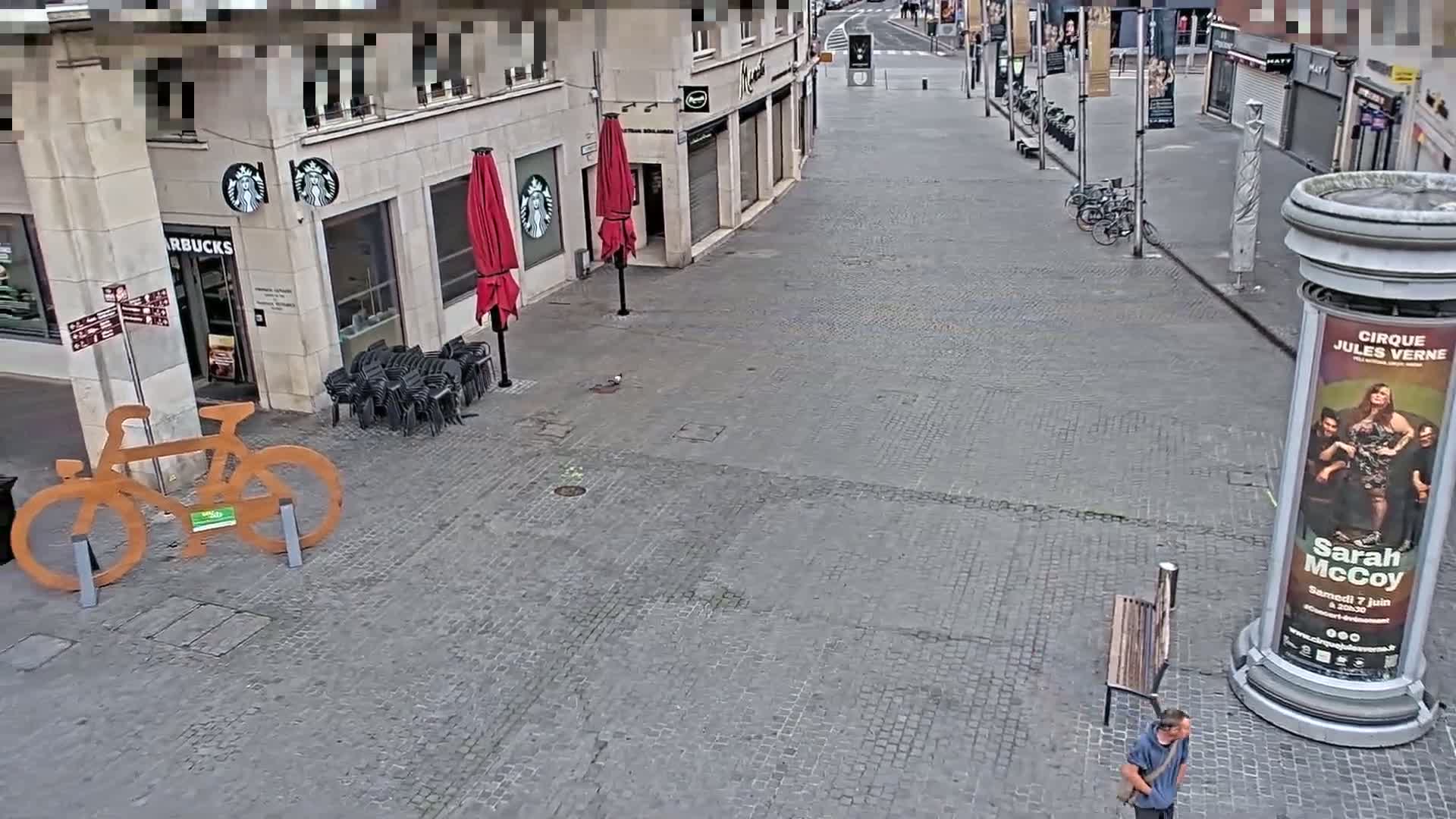 A mostly empty cobblestone plaza shows a Starbucks, several closed shops, a large wooden bicycle sculpture, red umbrellas, and a single person walking near a tall advertising column.