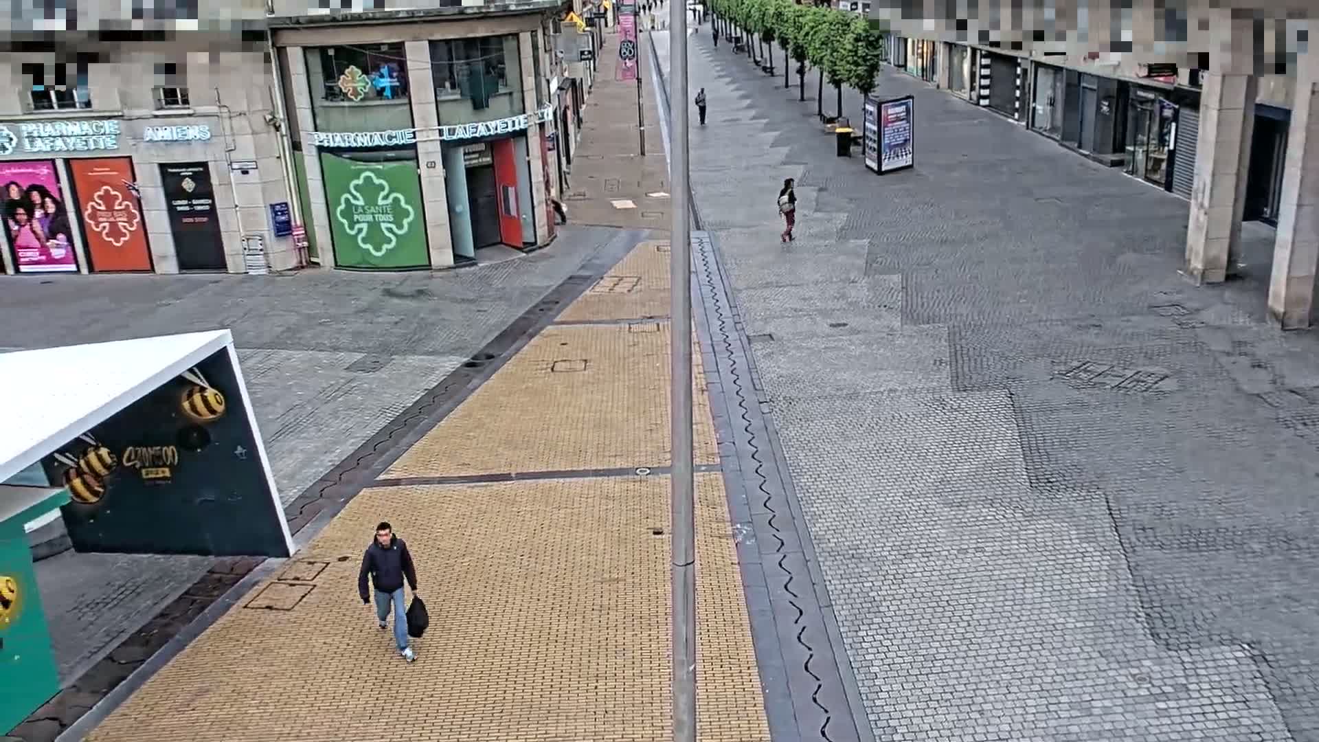 A mostly empty city square and street, paved with different colored stones, with a few pedestrians and buildings lining the street under an overcast sky.