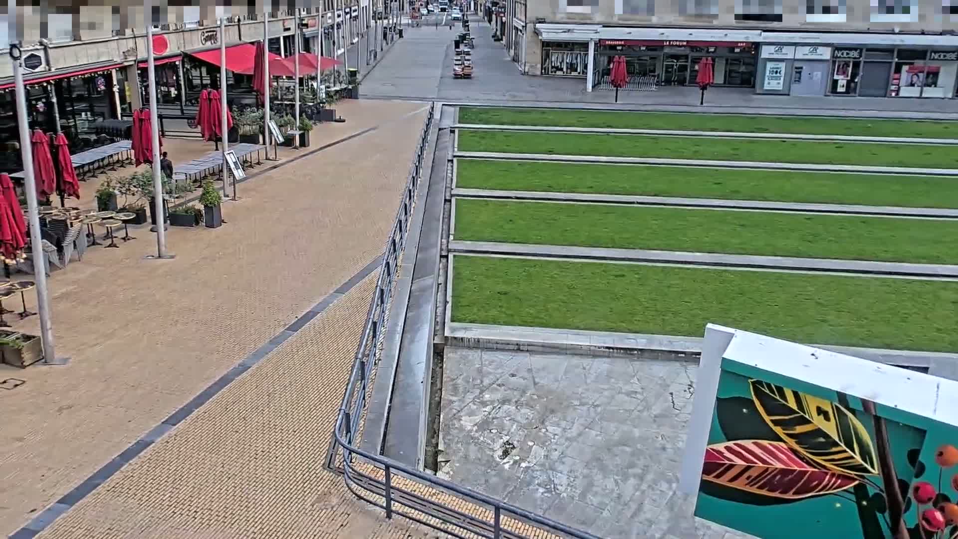 A paved plaza with red umbrellas shading outdoor cafe seating overlooks a sunken rectangular garden with neatly trimmed green grass, all under a clear sky.