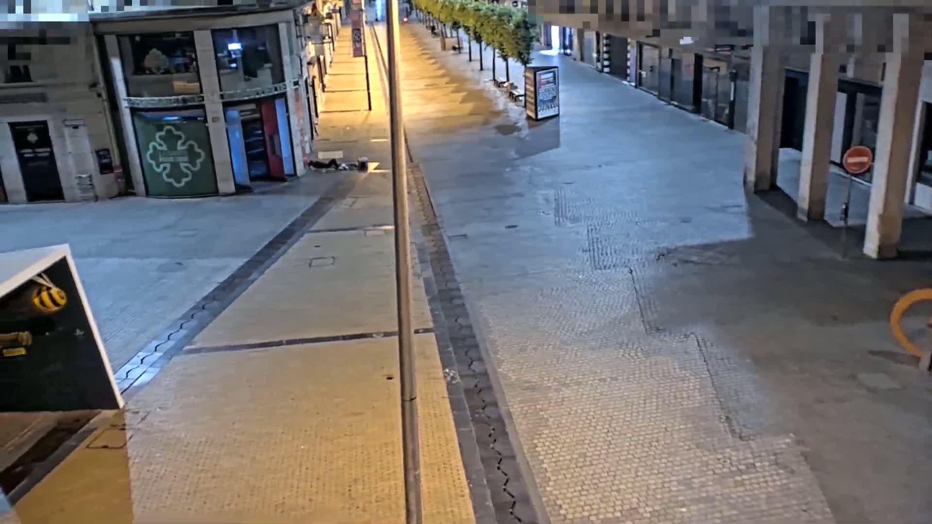 A mostly empty, paved street at night is lit by streetlights, with a person lying on the ground near a building.