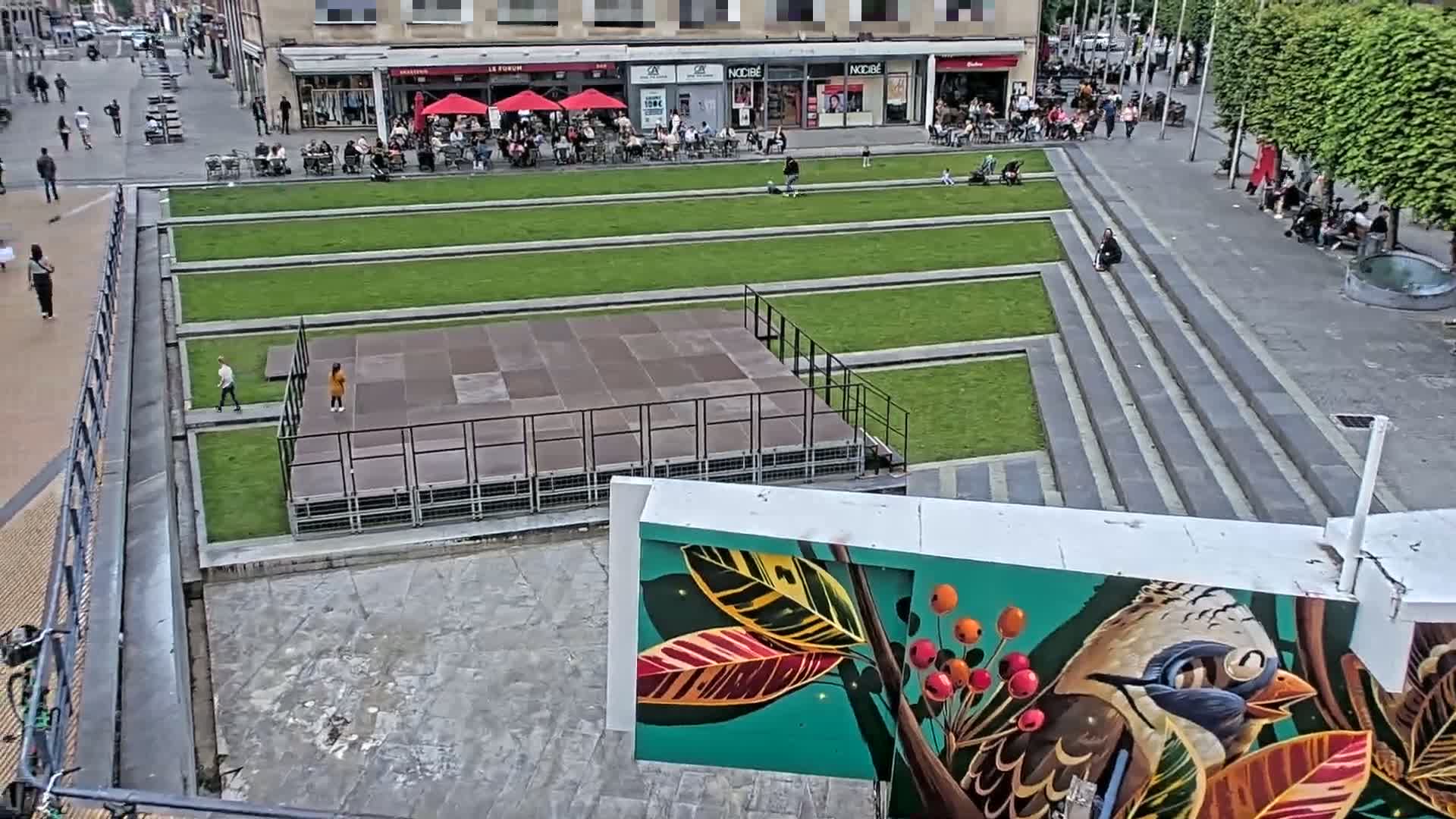 An outdoor urban plaza with a raised stage, grassy areas, and a mural of a bird and foliage is seen on a partly sunny day.