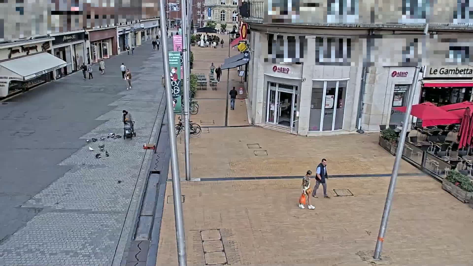A mostly empty pedestrianized street scene shows several people walking, a person feeding pigeons, and outdoor seating areas at a cafe, under an overcast sky.