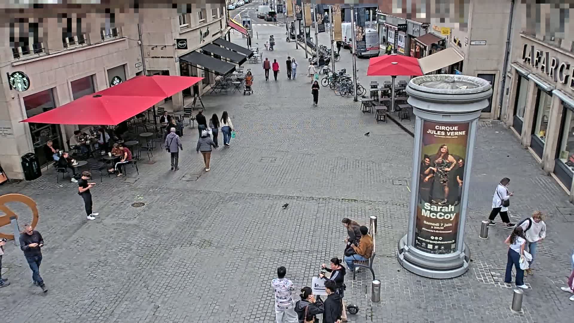 A cobblestone city square with people walking and sitting at outdoor cafes under red umbrellas on a partly cloudy day.