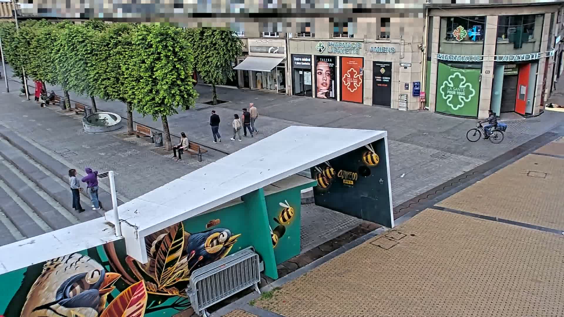 A city street scene on a partly sunny day shows several pedestrians and a bicyclist near shops and trees, with a large mural of birds and bees on a modern structure.