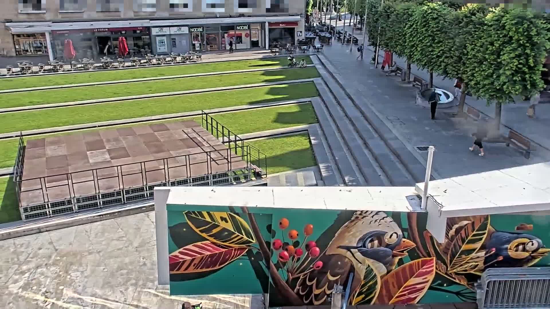 A sunny outdoor scene shows a paved plaza with a grassy area containing a raised platform, several pedestrians, and a large mural depicting birds on a branch.