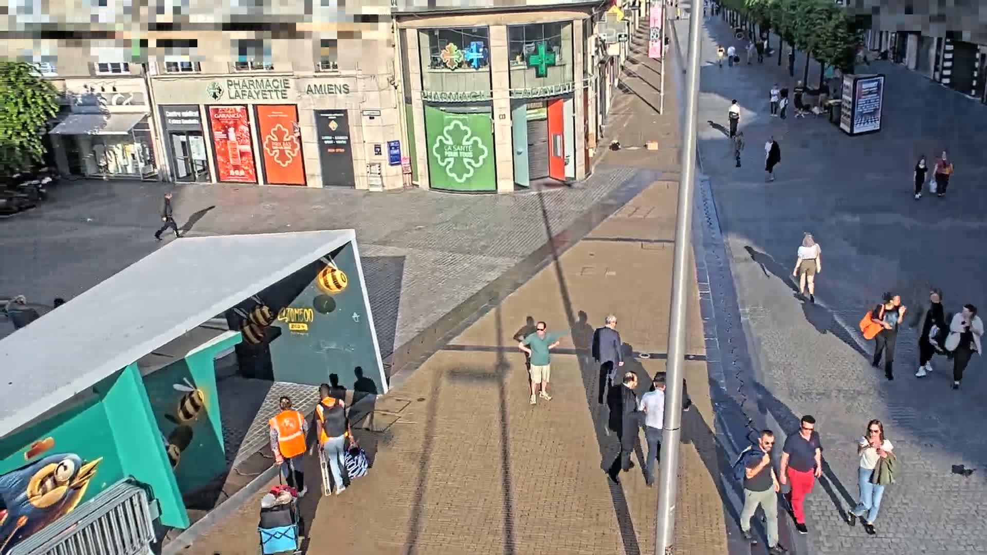 A sunny city square with pedestrians walking past buildings, one of which is a pharmacy, and a modern structure painted with bees.