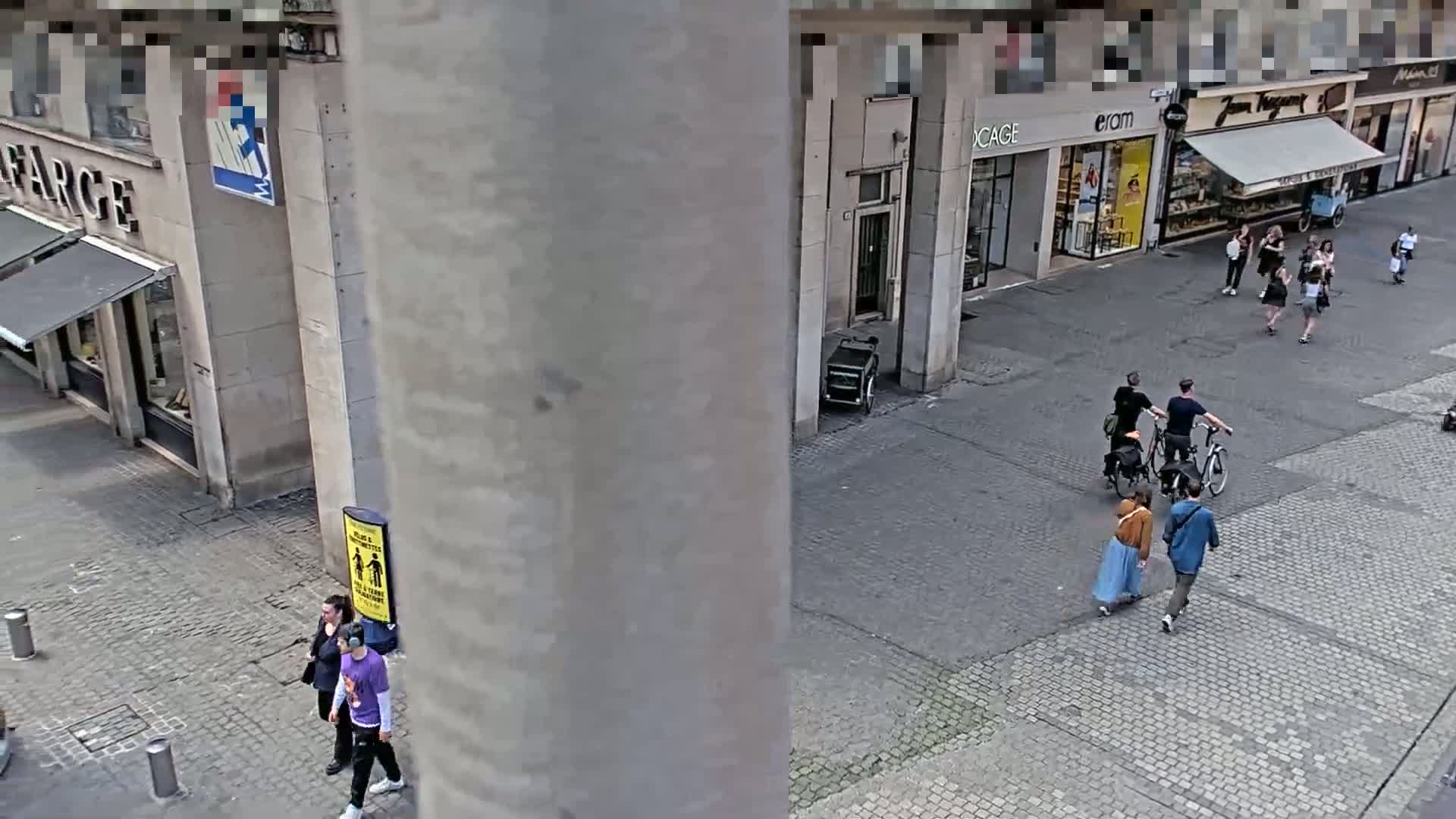 A high-angle view shows a cobblestone street with several pedestrians, some on bicycles, near shops under a clear sky.