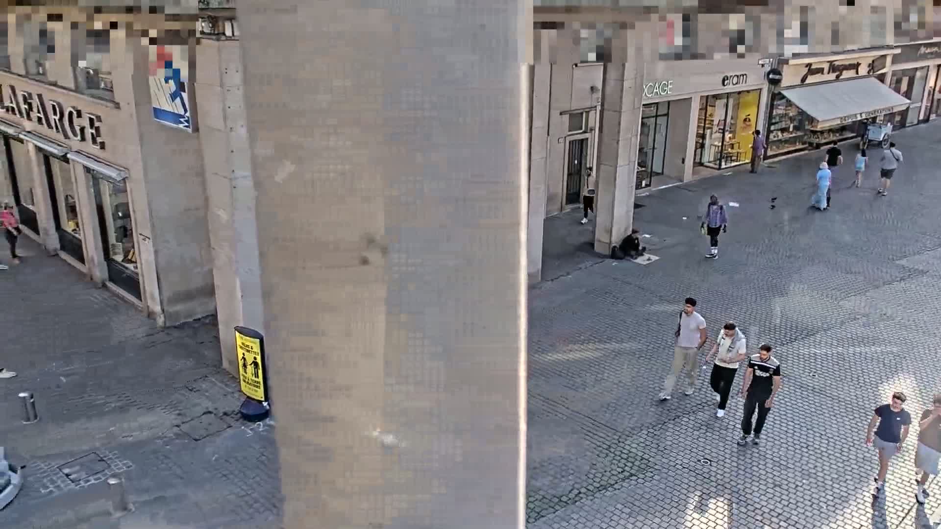 A high-angle view shows a cobblestone street scene on a sunny day with several pedestrians and buildings.