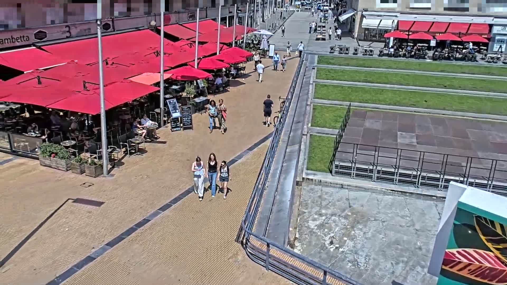 An outdoor scene on a sunny day shows a paved area with several restaurants under red umbrellas, people walking and sitting at tables, and a grassy area with a raised platform nearby.
