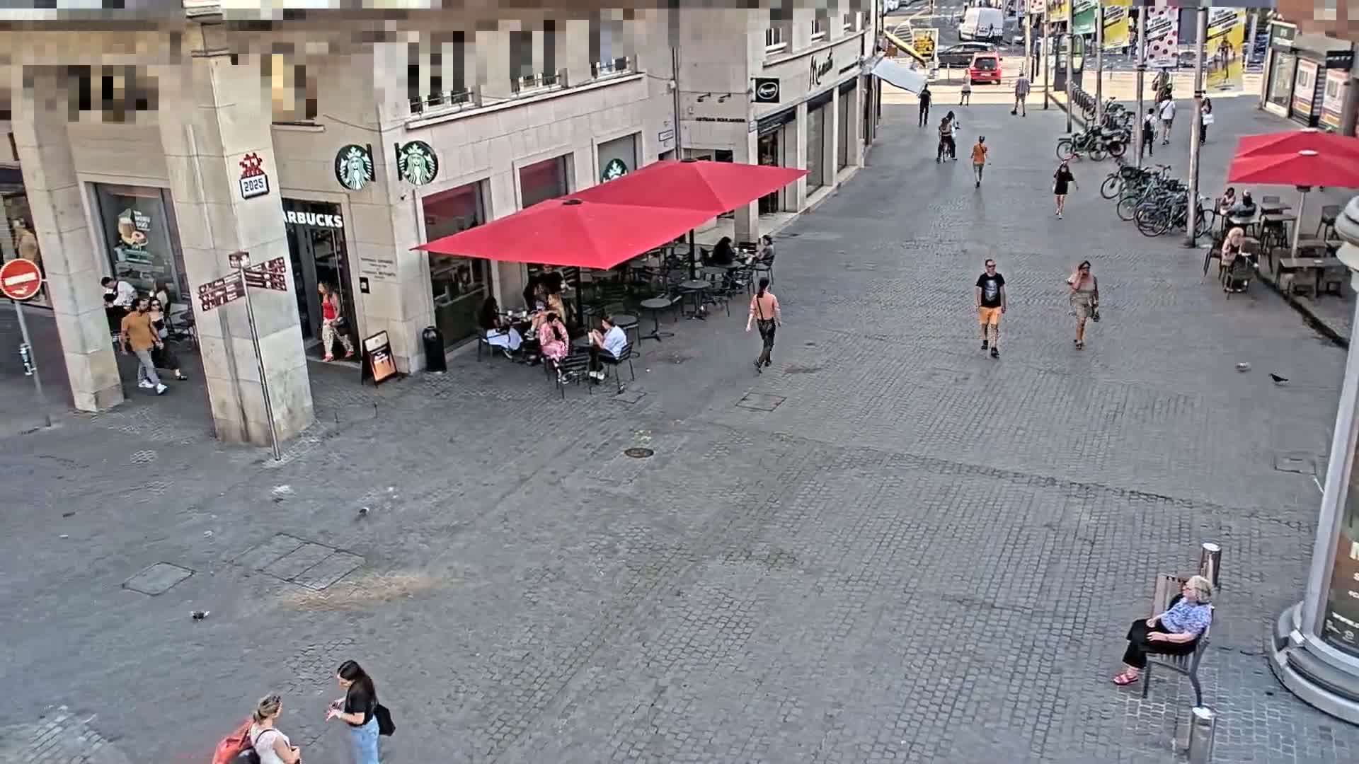 A paved outdoor area with several people walking and sitting at cafes under red umbrellas on a sunny day.
