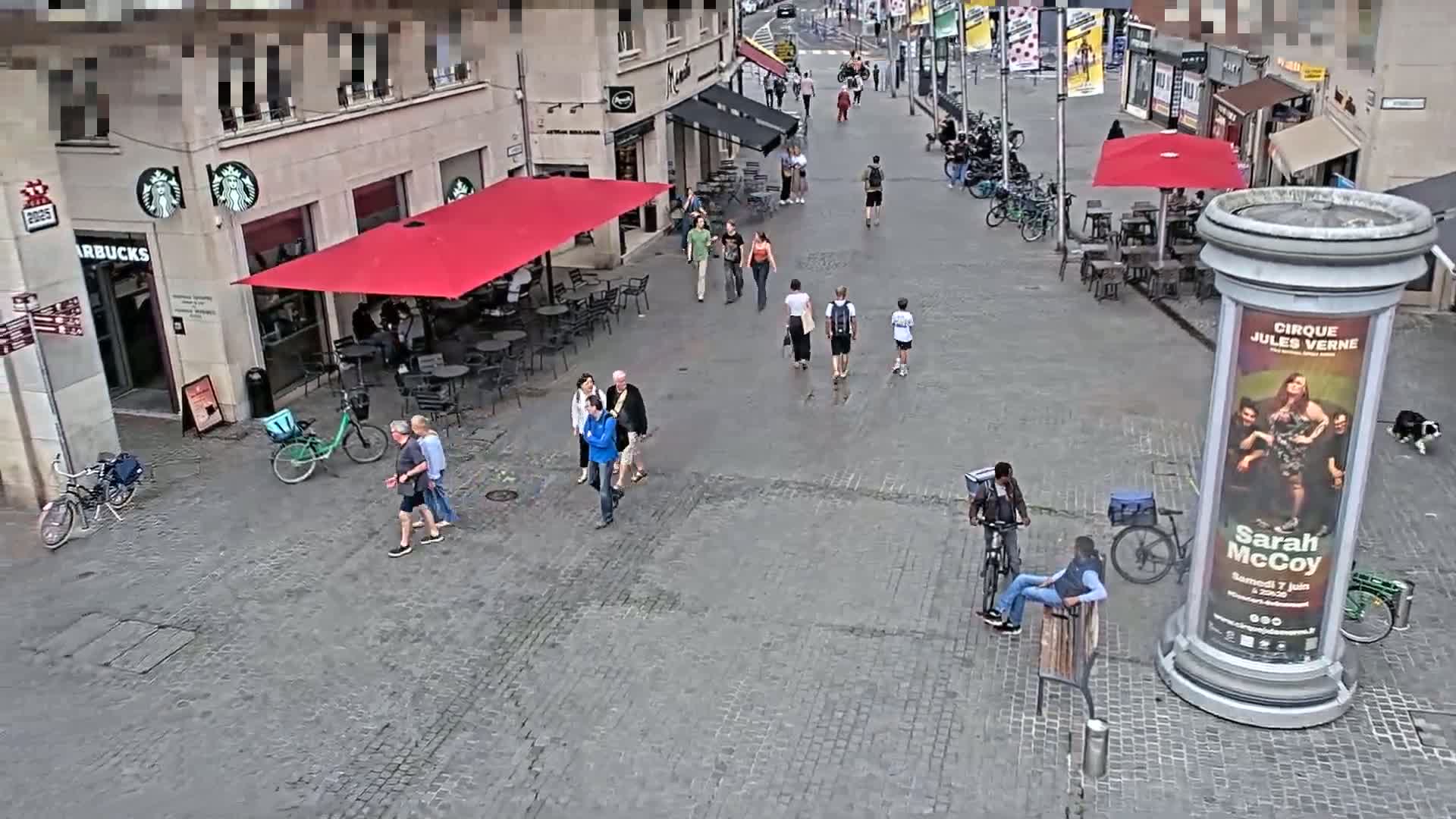 A cobblestone city square is populated with pedestrians, cyclists, and outdoor seating under red and black umbrellas, on a mostly clear day.
