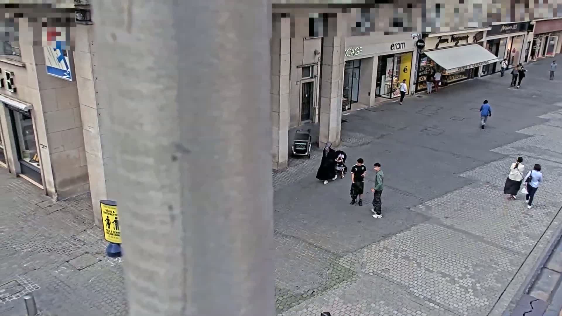 A high-angle view shows a cobblestone street with several pedestrians, including a person in a long black robe pushing a wheelchair, and two other people standing nearby.
