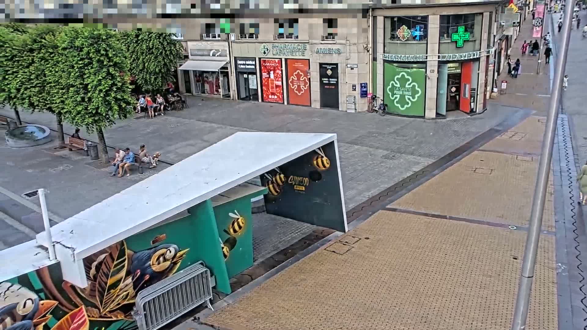A city square with people sitting on benches under trees, a pharmacy, and a modern structure with bee artwork on a sunny day.