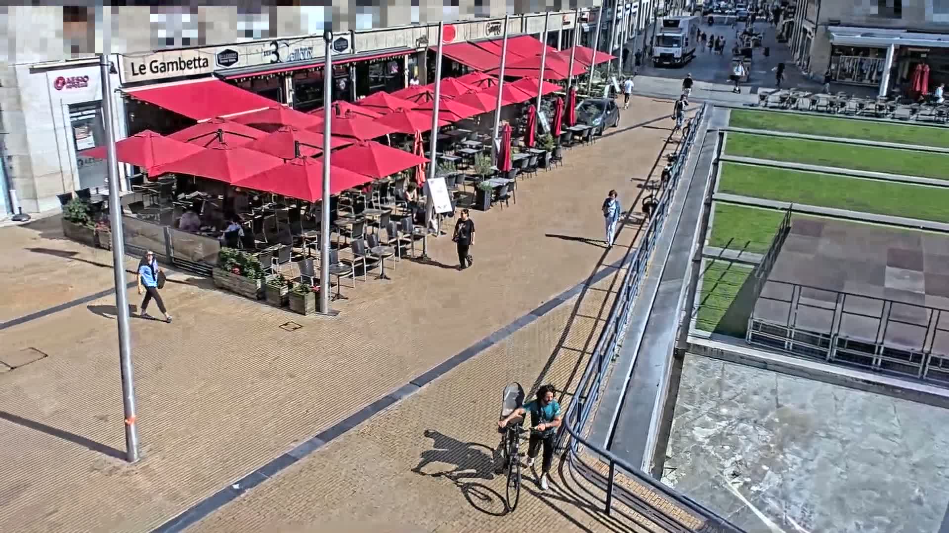 A person on a bicycle rides along a paved walkway next to a patio with red umbrellas and a grassy area on a sunny day.