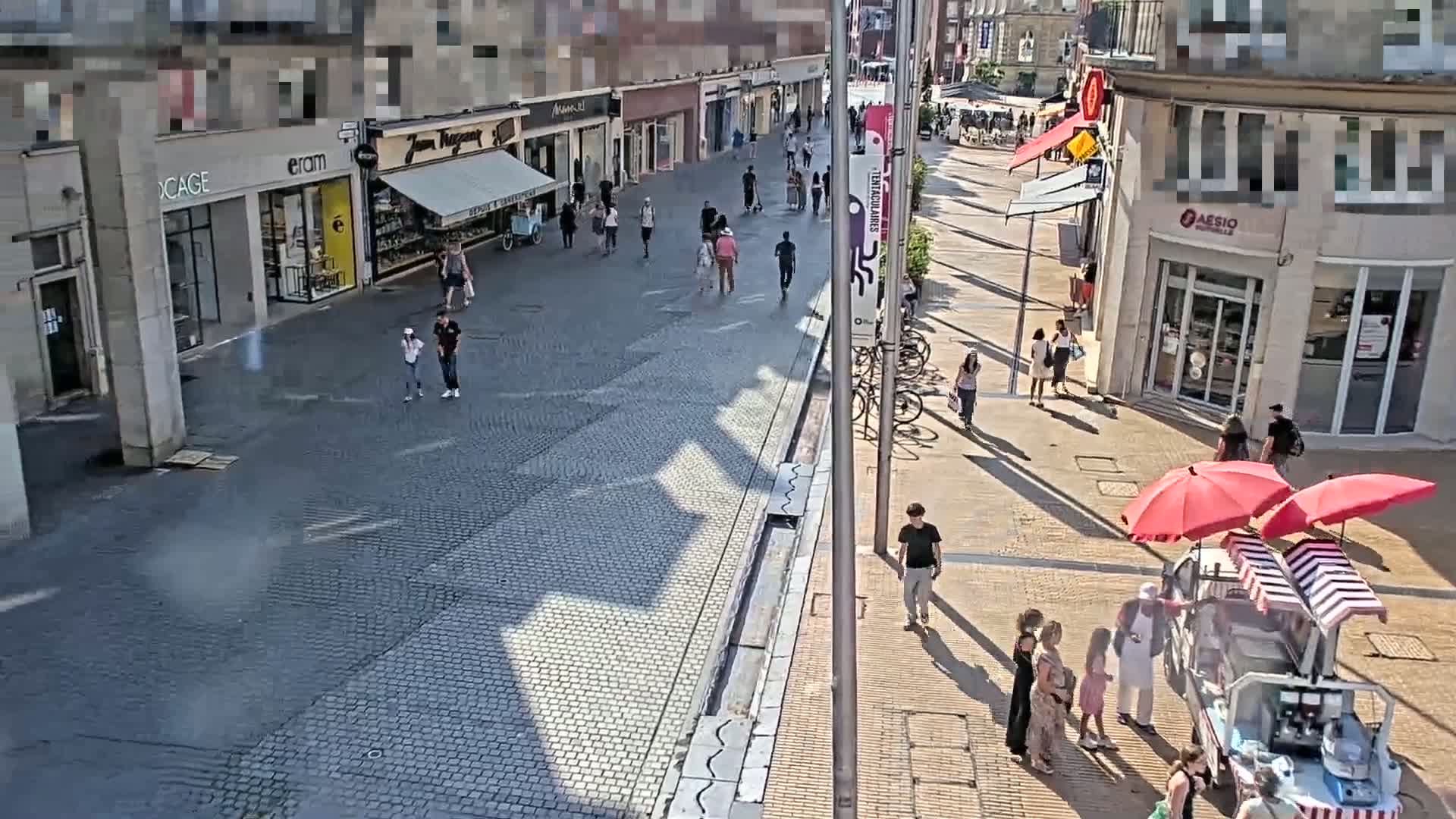 A paved pedestrian street with shops and people walking on a sunny day, and a food cart under red and white striped umbrellas.