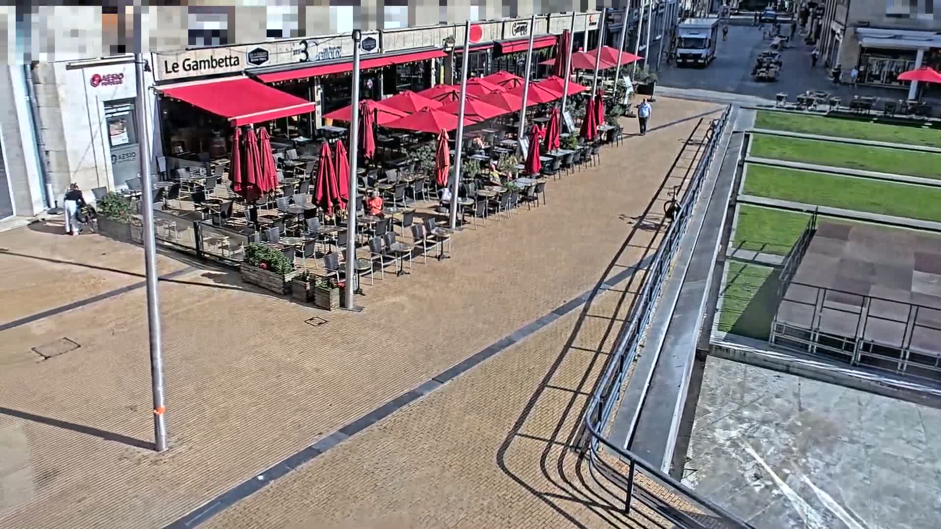 A sunny outdoor cafe with red umbrellas and patrons seated at tables, adjacent to a paved area with a railing overlooking a grassy expanse.
