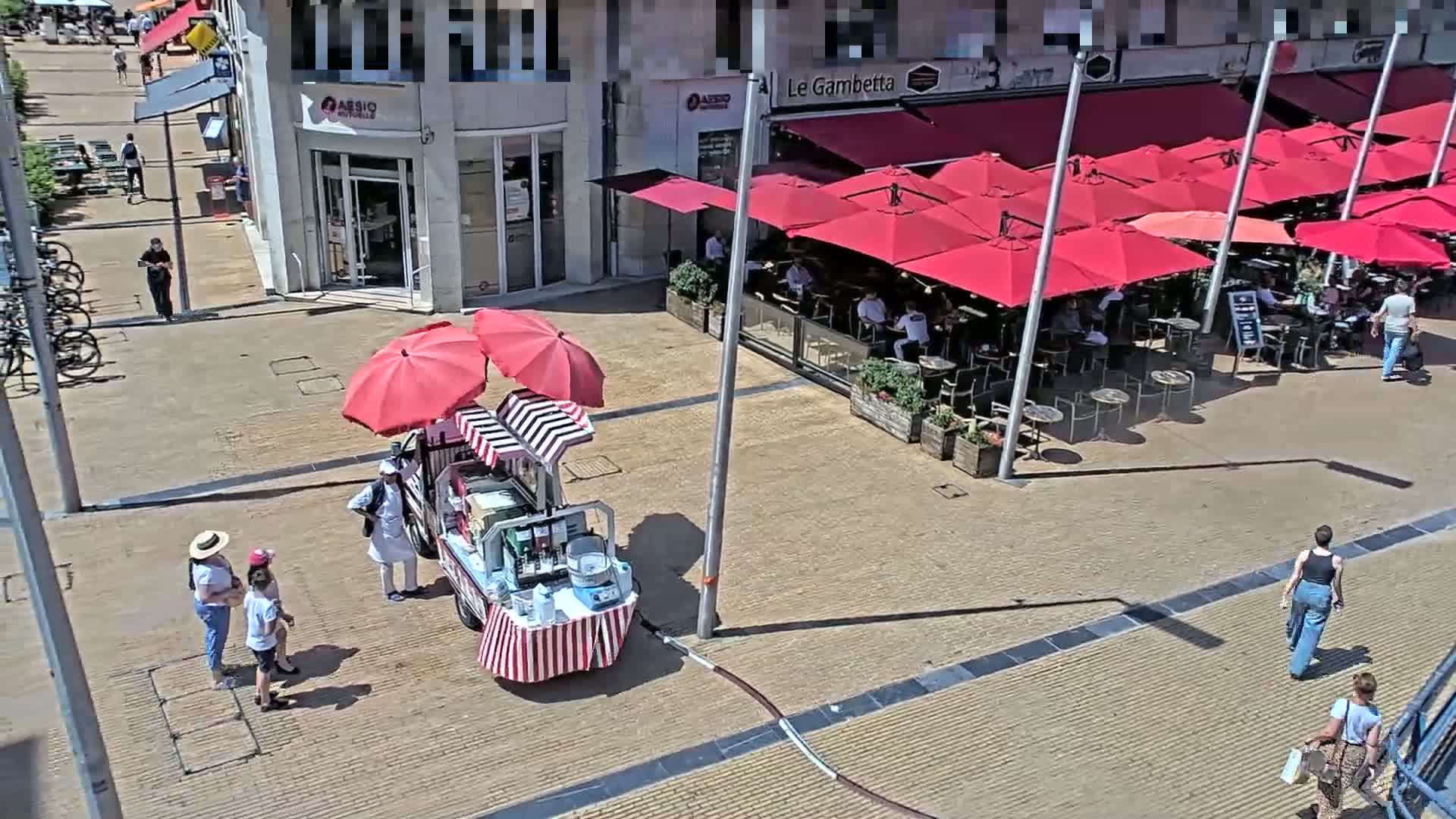 A sunny outdoor plaza scene shows a food cart with red and white striped canopy under two red umbrellas, several people milling about, and a cafe with numerous red umbrellas shading its outdoor seating area.