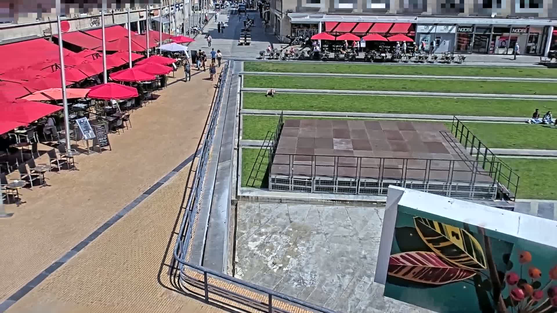 A sunny outdoor scene shows a paved area with red umbrellas shading cafe tables, a grassy park with a stage, and a building in the background.