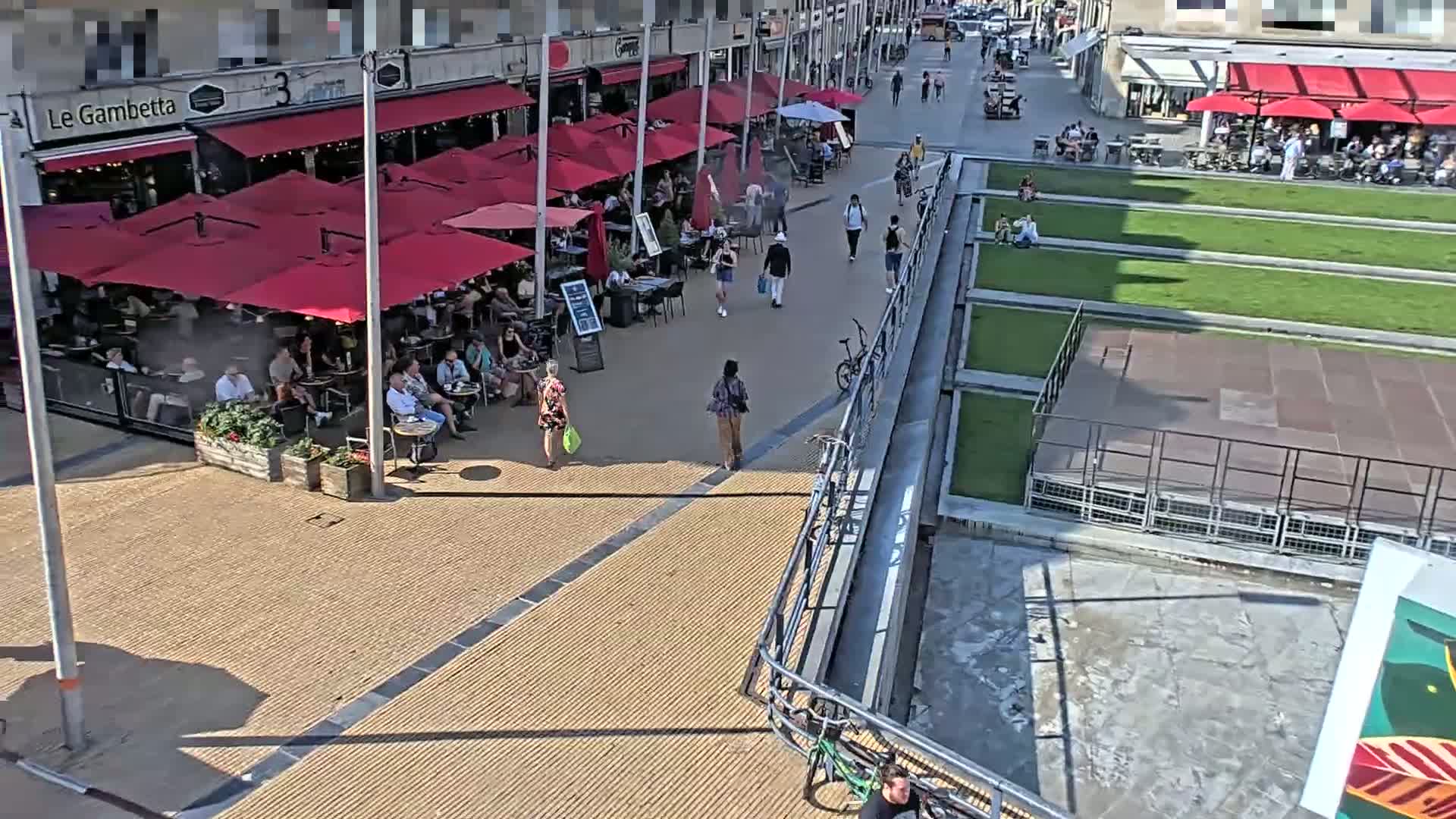 A sunny outdoor scene shows a paved pedestrian area with people walking and sitting at outdoor cafes under red umbrellas, adjacent to a grassy area.