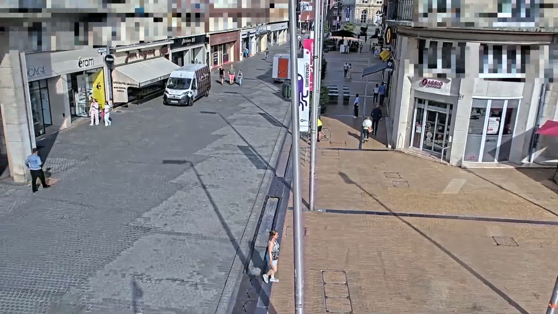 A sunny day shows a mostly empty paved street with several pedestrians and a cyclist near shops and buildings.