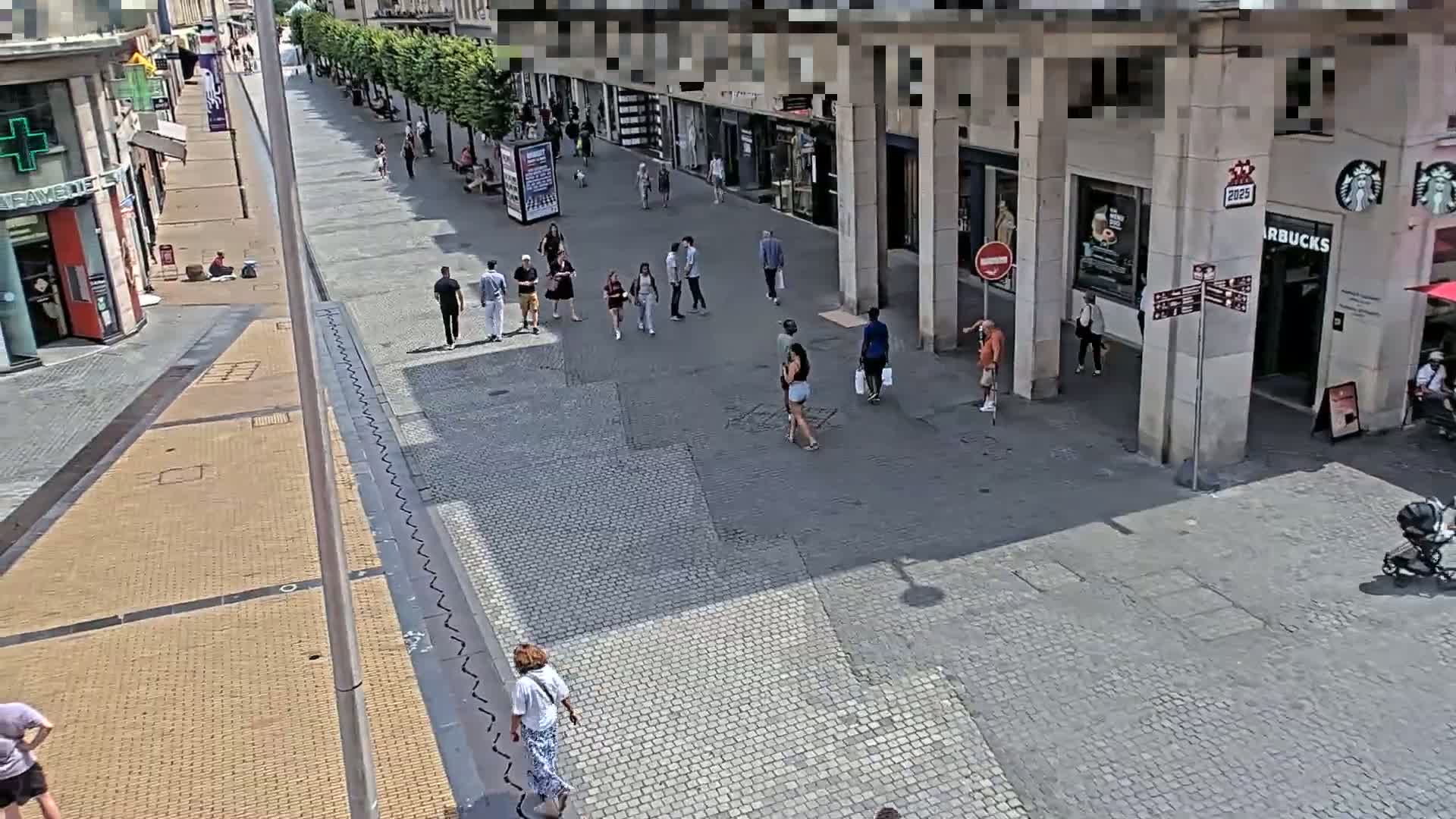 A sunny day on a paved pedestrian shopping street shows several people walking past shops and cafes.