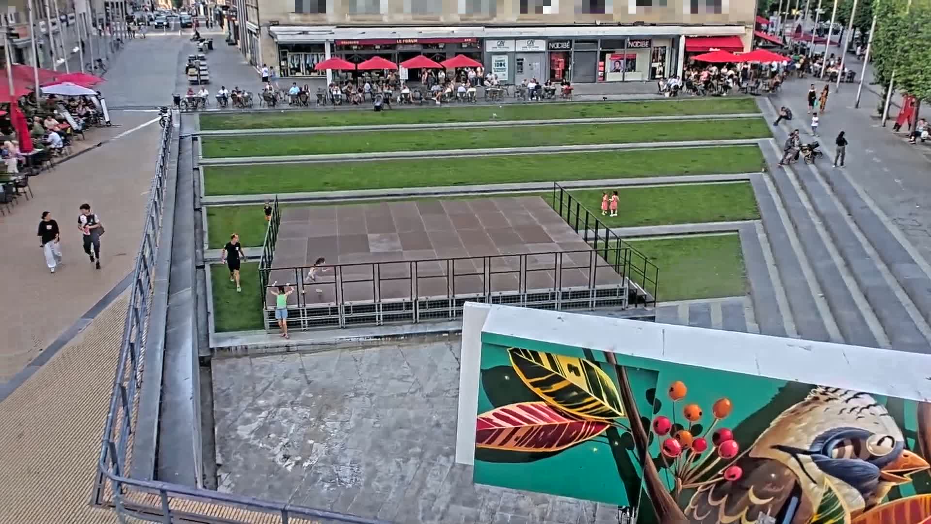 An elevated view shows a city square with a paved area, grassy sections, a stage, and people walking around on a sunny day.