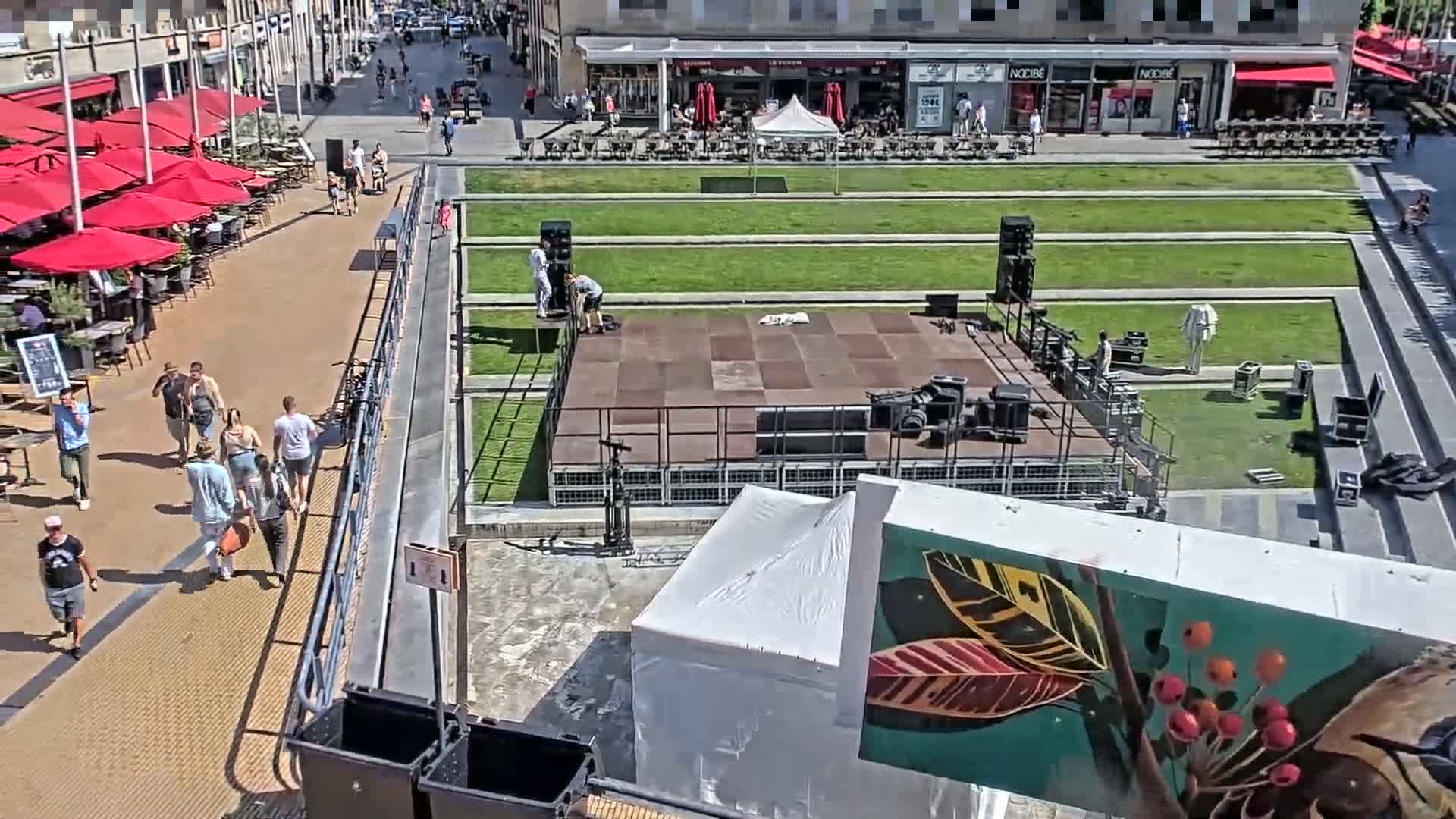 A sunny outdoor scene shows a paved walkway with people walking, outdoor seating under red umbrellas, and a large stage being set up in a grassy plaza.