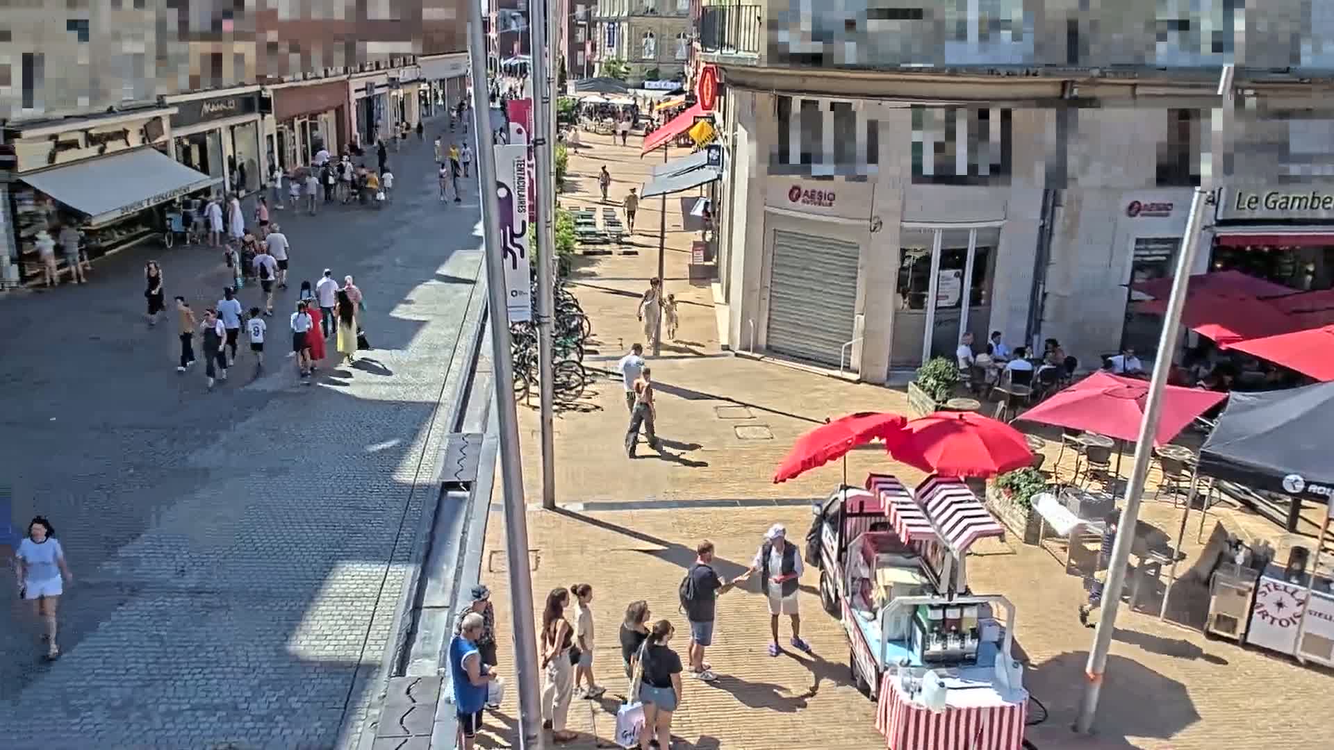 A sunny day on a paved pedestrian street shows numerous people walking and sitting at outdoor cafes under red umbrellas.